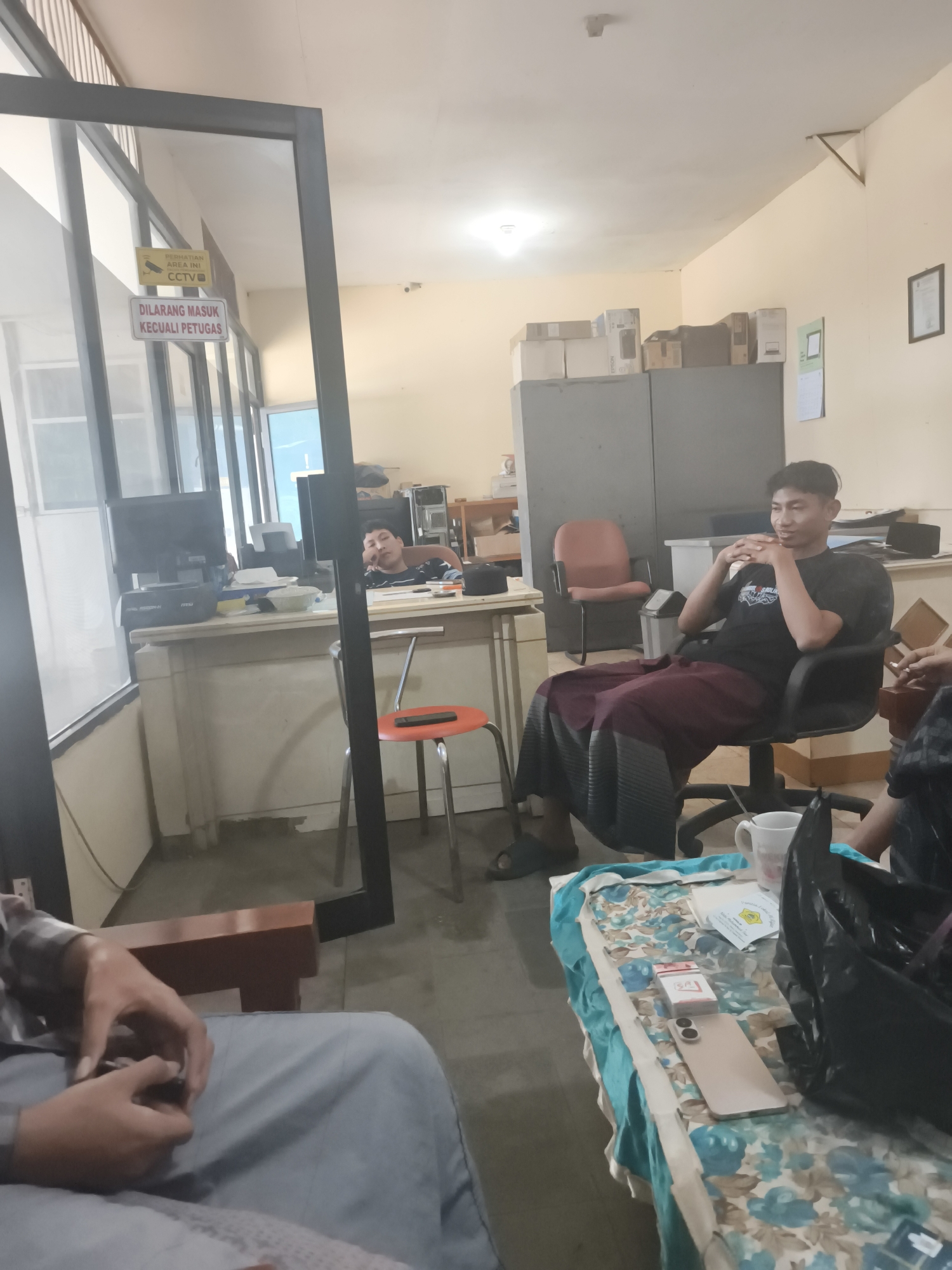 The image depicts an indoor scene, possibly an office or a waiting area, with several individuals present. In the foreground, a person is partially visible, wearing a grey checkered shirt and light blue pants, with their hands clasped in their lap. To the right, a young man is seated in a black office chair, wearing a black t-shirt and a maroon sarong. He is looking towards the camera with his hands clasped in front of him. Another person is seen reclining at a desk in the background, appearing to be asleep or resting, with their head propped on their hand.
The environment appears to be functional, with office furniture and equipment. There is a desk with computers and office supplies. A red chair with a black metal frame is placed in front of the desk. Various items are scattered on a table in the foreground, including a white mug, a pack of cigarettes, a mobile phone, and a black plastic bag. Signs are visible on a glass partition in the background, including one that reads "PERHATIAN AREA INI CCTV" and another with "DILARANG MASUK KECUALI PETUGAS". The walls are painted a light yellow, and fluorescent lighting is present on the ceiling. The overall impression is of a casual, possibly informal, gathering within an office setting. The presence of signs related to CCTV and restricted access suggests a controlled environment. Based on the signage, the location can be identified as Ketawang, Indonesia.