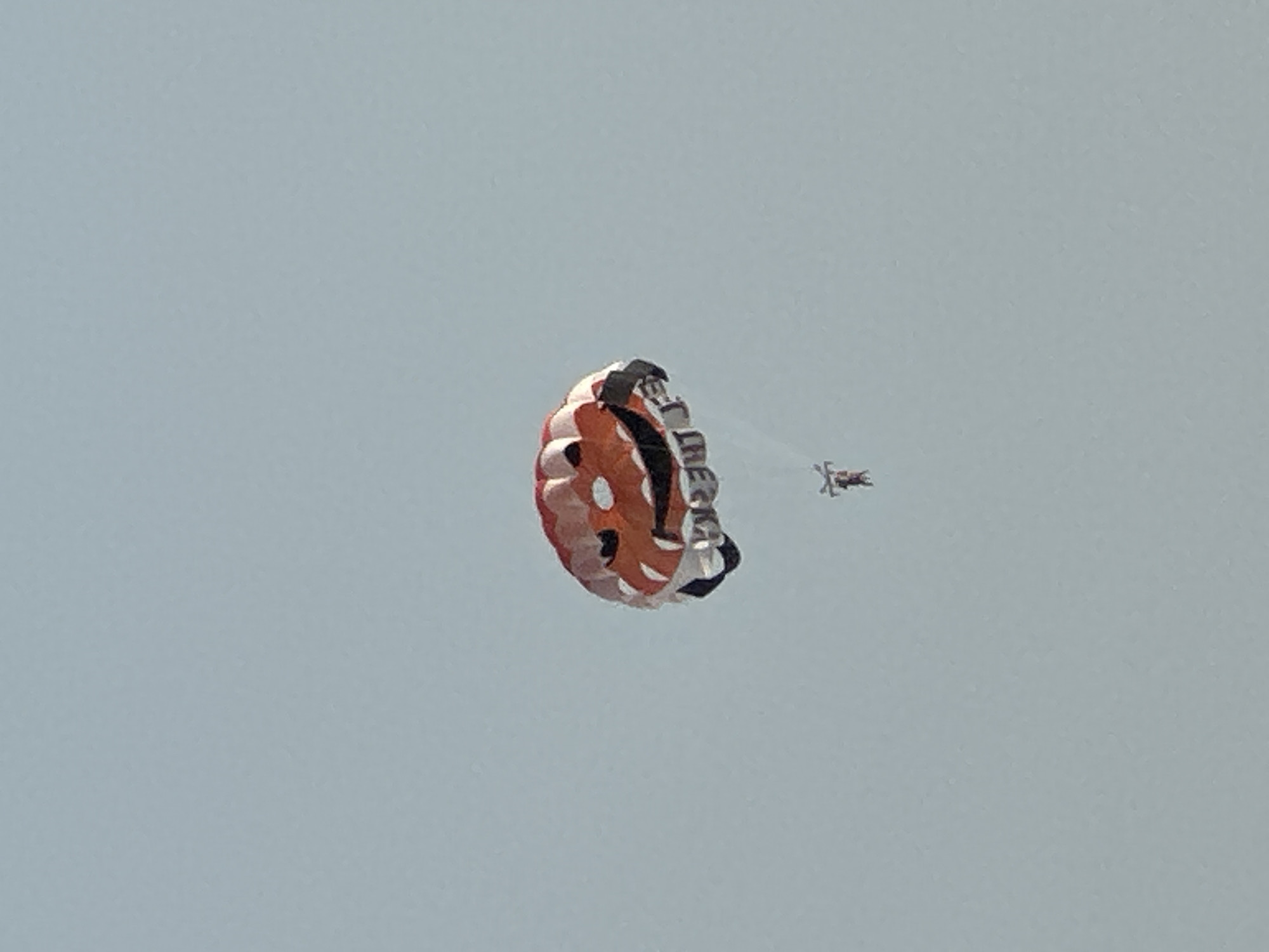 This media file captures a tandem parachute jump with a brightly colored parachute against a pale blue, clear sky, suggesting daytime with fair weather. The main subject is a large parachute, predominantly orange and white with black accents, unfurling in the air. The parachute appears to be a high-performance or artistic canopy, given its intricate design.
Visible on the parachute, though partially obscured and somewhat distorted due to the angle and fabric movement, are letters that appear to spell out "TIRE". This suggests a sponsorship or branding element related to a tire company.
Below and to the right of the main parachute, a second, much smaller parachute is visible. Attached to this smaller parachute is a skydiver in a dark suit, appearing as a small, silhouetted figure against the sky. This suggests a tandem jump is in progress, with one individual guiding the descent, or it could be two separate skydivers.
The scene evokes a sense of adventure and freedom, typical of skydiving activities. There are no specific environmental cues to pinpoint the exact location within Boreti, Montenegro, beyond it being an outdoor skydiving area. The overall impression is one of a successful and visually striking aerial maneuver.