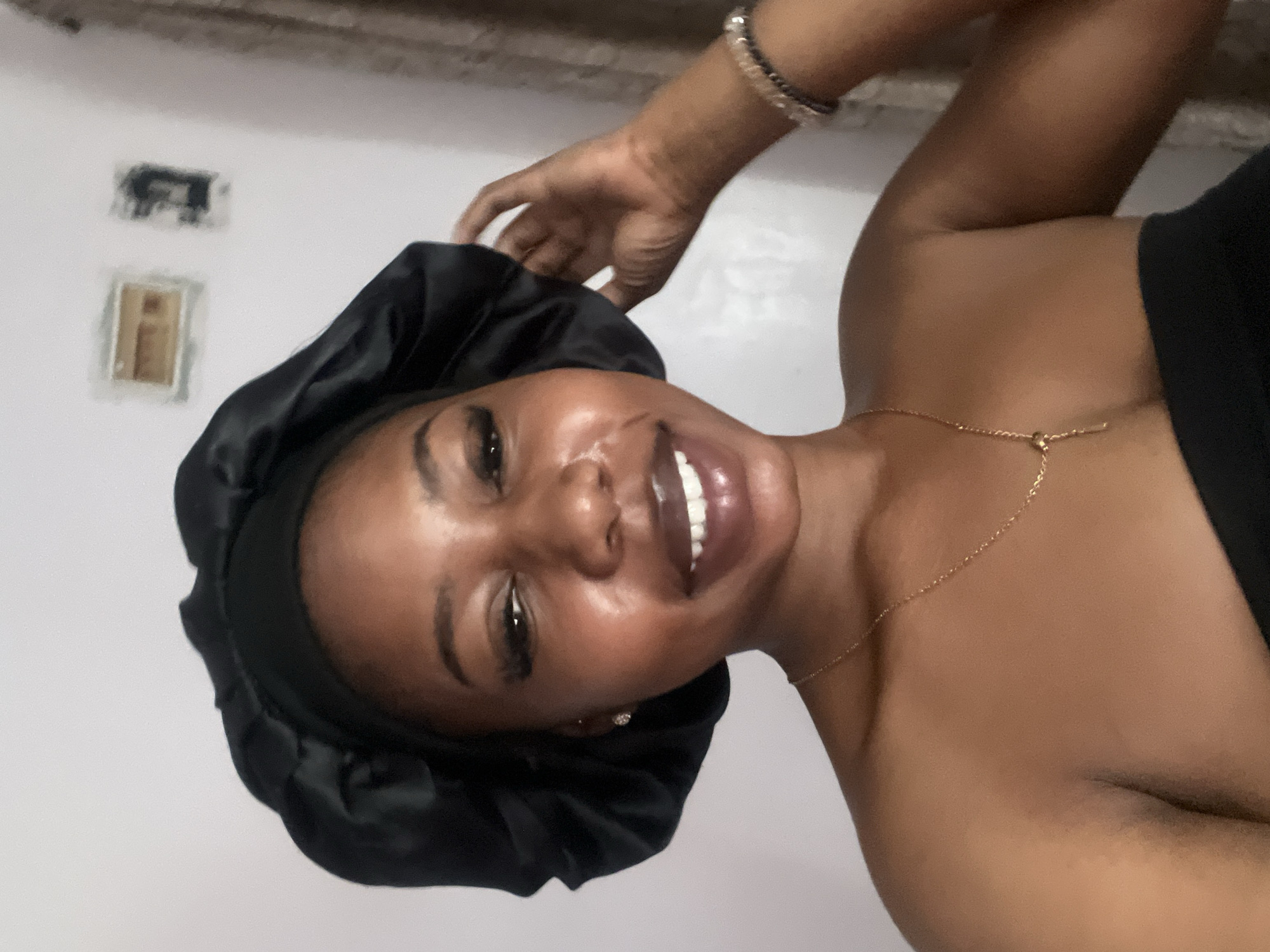 A young Black woman is captured in a selfie, looking towards the camera. She is wearing a black satin bonnet that covers her hair. Her makeup includes dramatic eyelashes and lip gloss. She is also wearing a delicate gold necklace with a pendant and a beaded bracelet on her right wrist. The background appears to be an indoor setting, possibly a bedroom, with a light-colored wall that has some indistinct items mounted on it, including what look like light switches or electrical outlets. A curtain is visible on the right side of the frame. The lighting suggests it may be daytime, with light illuminating the woman's face and shoulders. The overall impression is a casual, personal moment. There is no visible indication of the location being in Jos, Nigeria.