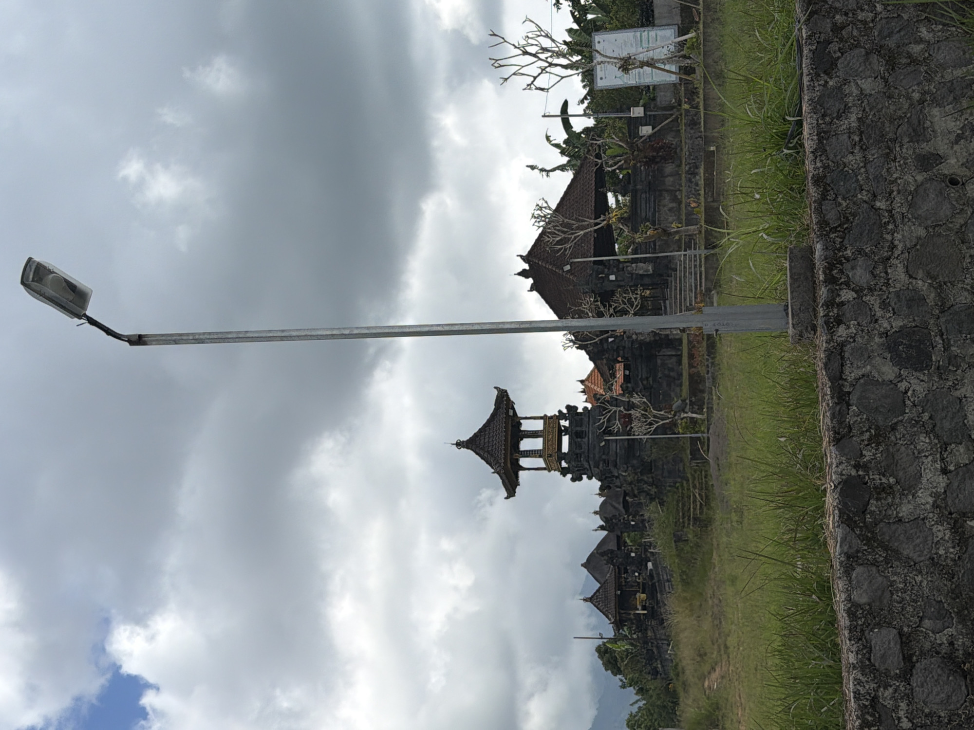 The image shows a street lamp post in the foreground, obscuring a portion of a traditional Balinese temple complex. The temple features several multi-tiered roofs and stone structures, characteristic of the region. Lush green grass covers the grounds around the temple, and in the background, distant mountains are visible under a cloudy sky. A stone wall runs along the bottom of the frame. This scene is located in Kintamani, Indonesia.