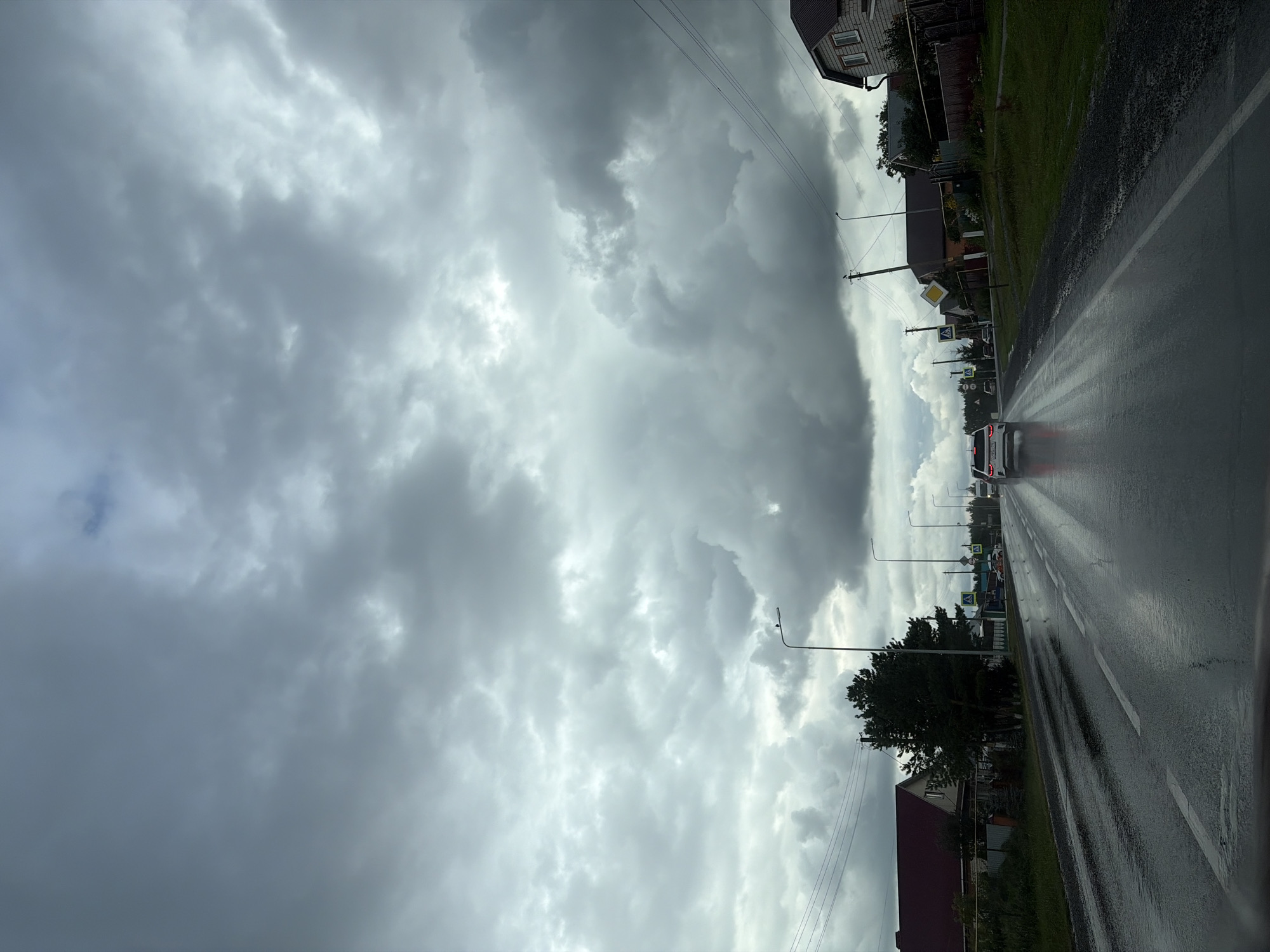The image depicts a view from a vehicle driving on a wet, asphalt road in a rural or suburban area. The sky is overcast with dramatic, dark gray clouds. On either side of the road, there are houses with various roof colors, fences, trees, and streetlights. There are also power lines visible. A white car is driving ahead on the road, and its taillights are illuminated. The road surface reflects the sky and the surrounding environment due to the wet conditions. There are crosswalk signs present, indicating potential pedestrian crossings. The overall impression is of a cloudy, potentially rainy day.