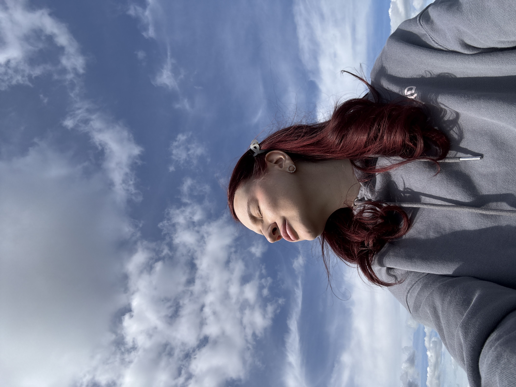 A woman with long, auburn hair, partially tied back with a small, translucent clip, stands outside under a partly cloudy sky. She wears a gray hooded sweatshirt and round earrings. Her eyes are closed, and her face is turned toward the sky. The image is shot from a low angle, looking upward. There is no verifiable information to confirm the location is Bol'shoye Kozino, Russia, and no other objects, actions, or interactions are depicted in the image.