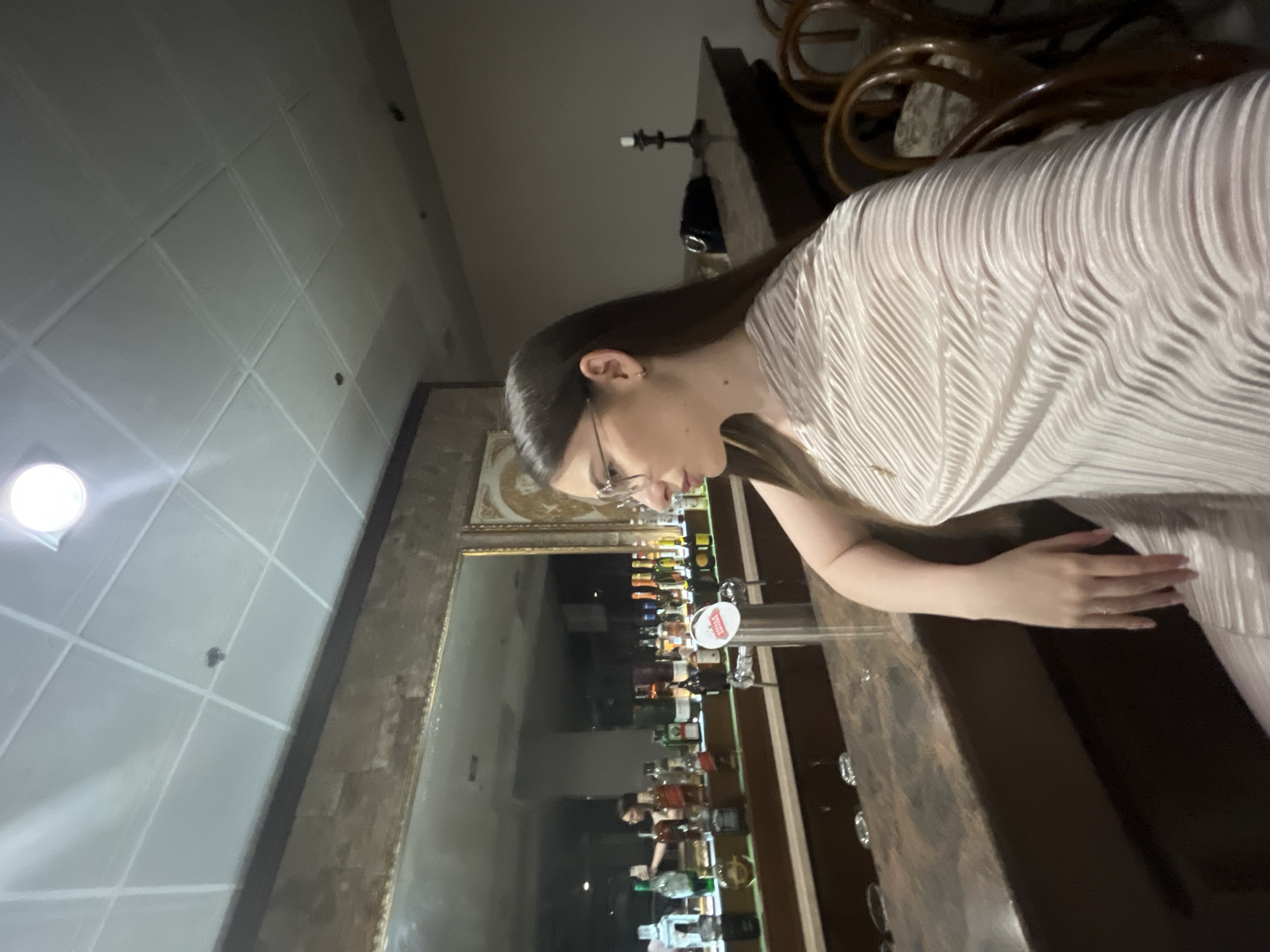 The image shows a young woman wearing glasses and a white pleated dress standing in front of a bar in an unknown location. The bar has a mirror behind it, reflecting a selection of alcoholic beverages. The woman's hand is resting on the bar's surface. Overhead, the ceiling is tiled with a recessed light fixture. A portion of the bar's ornate wooden structure is visible in the upper right corner.