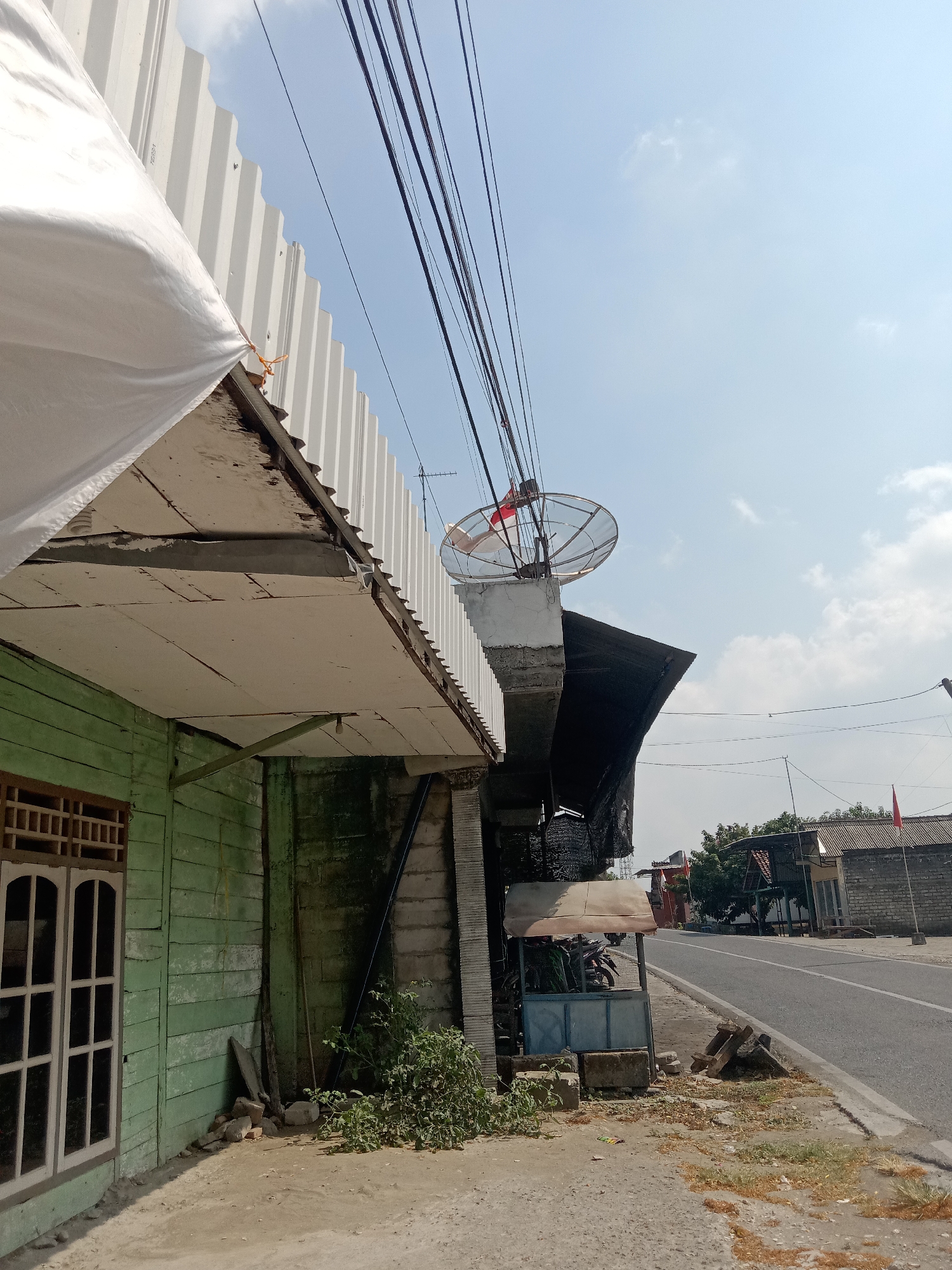 In Siwalan, Indonesia, a satellite dish mounted on a concrete structure is visible against a bright, partly cloudy sky. The dish is positioned above a section of corrugated metal roofing and appears to have a red and white Indonesian flag attached to it. Adjacent to the satellite dish, a green wooden building with a multi-pane window is partially visible on the left side of the frame. The building's exterior shows signs of weathering. In the foreground, dirt and scattered debris are present on the ground. Further into the background, a paved road runs alongside several buildings with various roofing materials. One of these buildings has a small, open-air kiosk in front of it, sheltering a motorcycle. Red flags are also visible on poles in the distance. Overhead, a cluster of dark electrical wires spans across the scene.