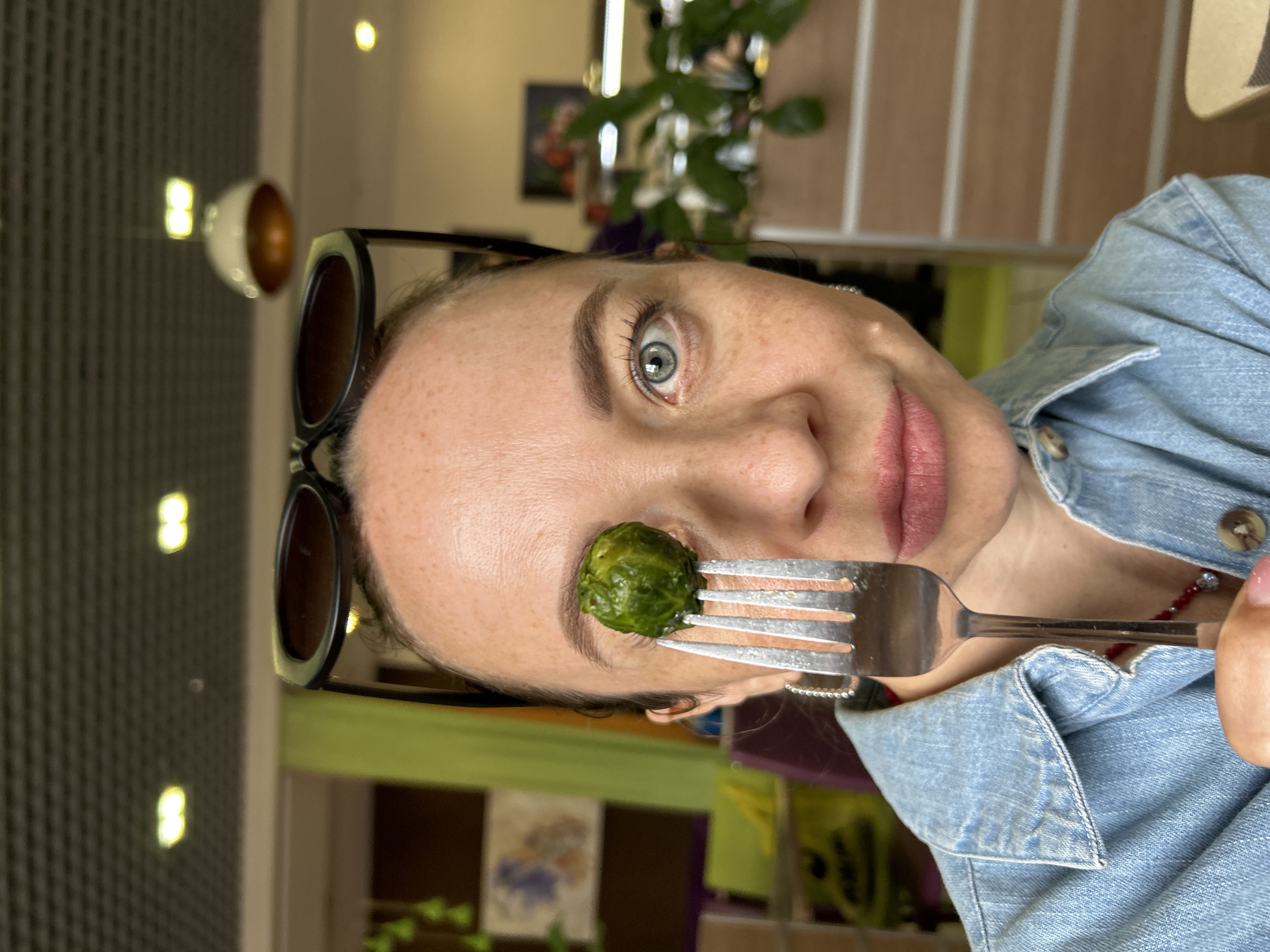 A close-up, sideways portrait shows a young woman with light skin and blue-green eyes, wearing a light blue denim shirt. She is holding a fork with a single Brussels sprout impaled on it, positioned in front of her right eye. The woman has freckles across her nose and cheeks and is wearing dark-rimmed sunglasses pushed up onto her forehead. Her lips are slightly parted and appear to be lightly colored with lipstick. A thin red beaded necklace is visible around her neck.
The background is blurred, suggesting an indoor setting. The lighting is warm, with some bright reflections visible on the left side of the image, possibly from overhead lights or windows. The overall impression is that of a casual and playful moment captured during the day. No visible text is present in the image.