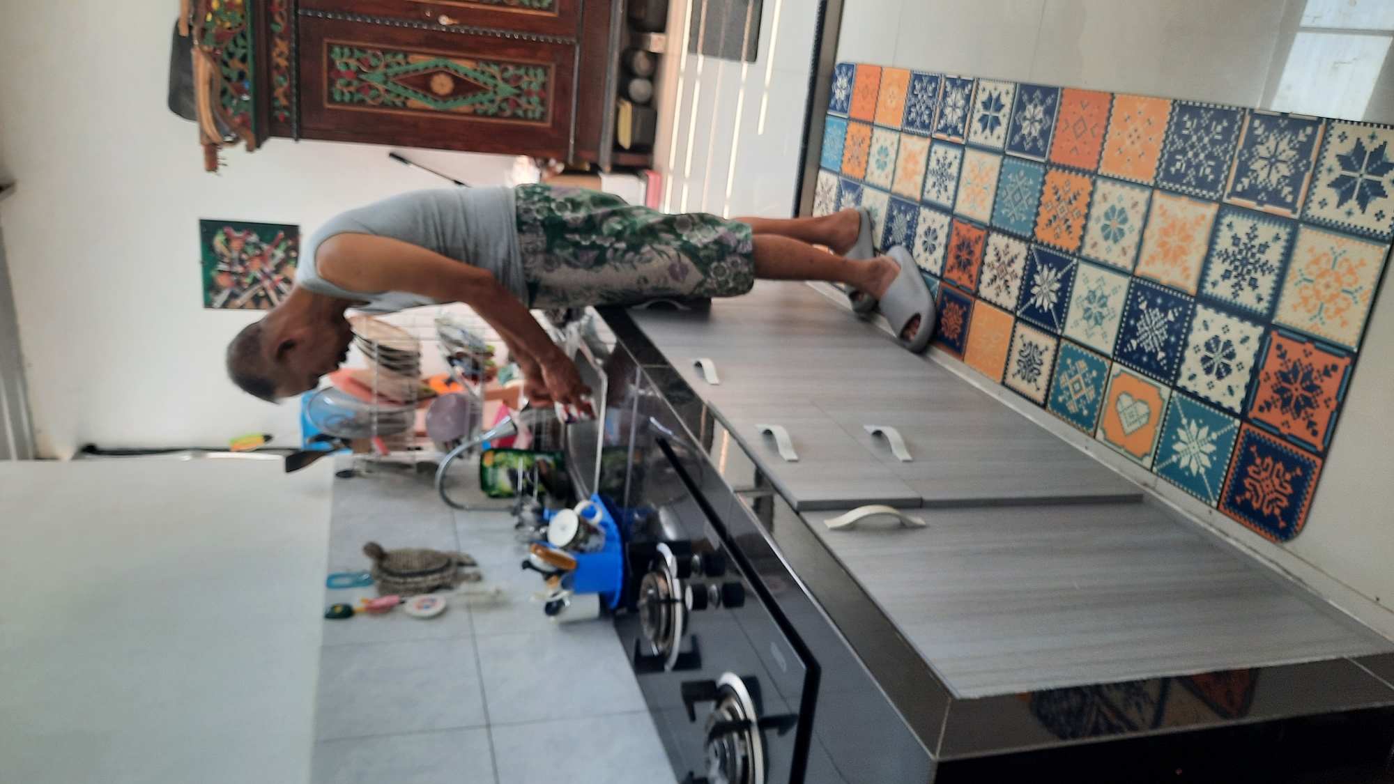 In a kitchen setting, an adult male with short, dark hair and a mustache, wearing a grey sleeveless shirt and patterned shorts, stands at a sink and appears to be washing dishes. The kitchen features a black countertop with a built-in stovetop, grey cabinetry below the counter, and a tiled backsplash. A colorful rug with a mosaic tile pattern is laid out on the floor in front of the cabinets. In the background, a dark wooden cabinet with ornate carvings is visible, along with a piece of artwork on the wall. The environment suggests a domestic space, likely a home kitchen, located in Salatiga, Indonesia.