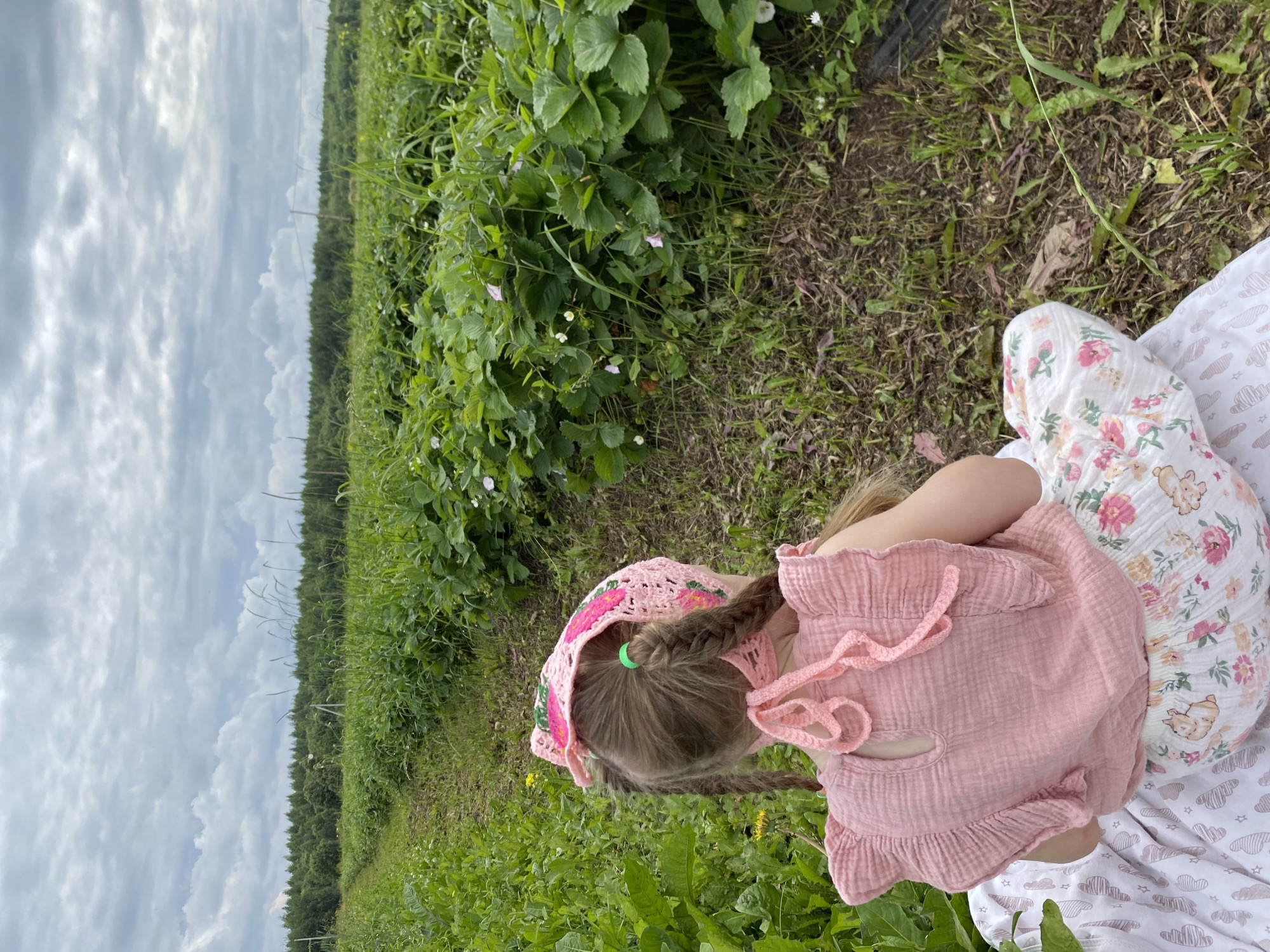 The image shows a young girl sitting in a field of green plants, likely a strawberry patch, in Foteni, Russia. She is wearing a pink crochet hat with flower designs and a light pink ruffled top. Her blonde hair is in two braids, tied with a green hair elastic. Her legs are crossed, and she is wearing patterned pajama bottoms with clouds and bears.
The setting is an outdoor field with abundant green foliage, suggesting a natural or agricultural environment. In the background, a dense line of trees creates a horizon. The sky is partly cloudy with some white and gray clouds. The overall impression is that of a peaceful, perhaps summer day, with the girl enjoying the outdoors. There is no visible text in the image.
