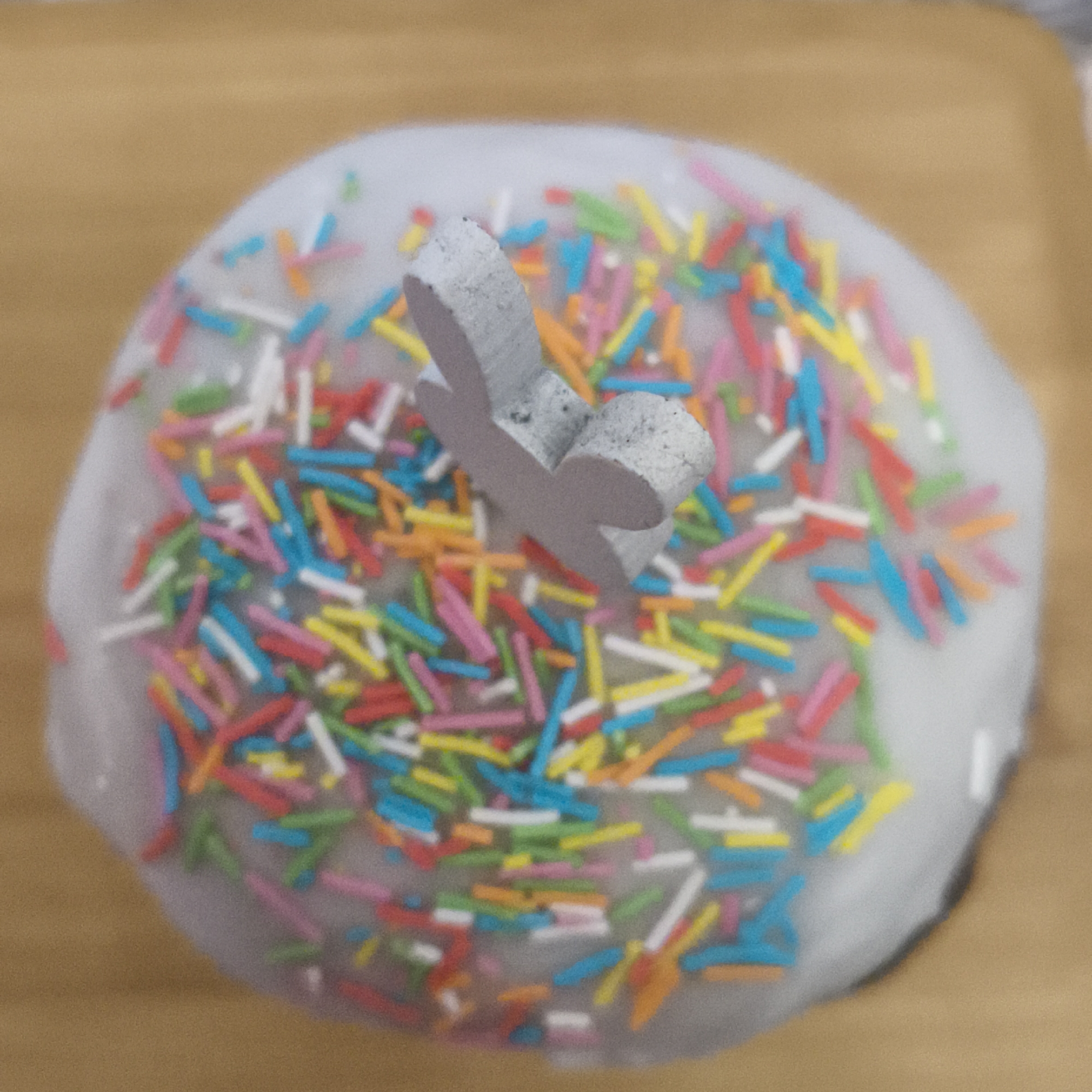 A close-up overhead shot depicts a frosted Easter cake. The cake is decorated with multicolored sprinkles and a small, gray, bunny-shaped adornment on top. The cake rests on a light brown wooden surface.