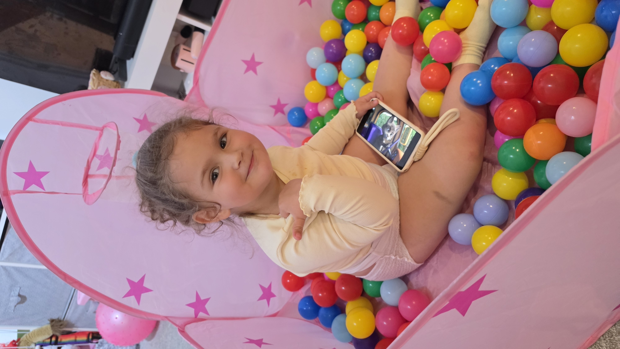 A young child lies in a pink, star-decorated ball pit filled with multicolored plastic balls. The child is wearing a light-colored, long-sleeved onesie with white socks and is holding a smartphone, appearing to watch something on it. The child is smiling and gesturing with their free hand. The background shows part of a room with shelving. The location is a private residence, not the city of London, United Kingdom.
