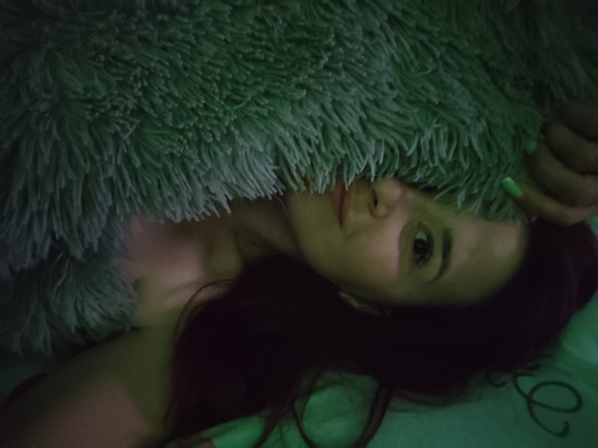 A woman is lying down and partially covered by a furry, green blanket. Her head is slightly tilted. She has dark hair. Her nails are painted green. She appears to be indoors, lying on a green surface, potentially a bed. The image is taken at a close range. No specific details are apparent that would identify the location as Фоки, Russia.