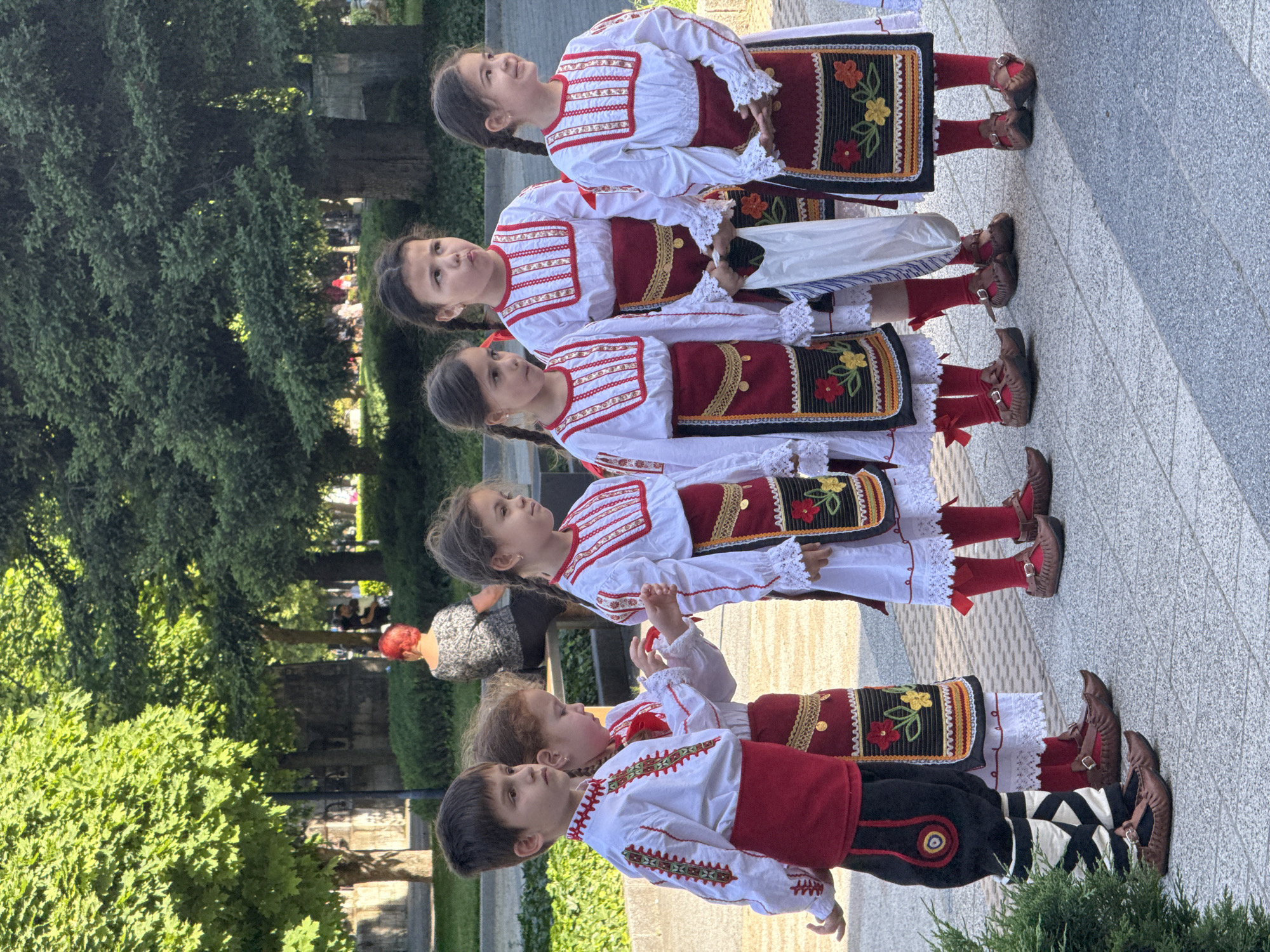 A group of five children, dressed in traditional Bulgarian folk costumes, stand in a line in what appears to be a park in Pleven, Bulgaria. The children are in ascending order of height from front to back. The costumes feature white shirts with red and white embroidery, red sashes or waistcoats, and black skirts or pants with embroidered detailing. The girls have their hair braided. All children wear red socks and brown leather shoes or boots. The foreground is a paved area, and in the background, there are trees, greenery, and buildings that might be part of the urban landscape of Pleven. Some blurred figures of other people can be seen further back.