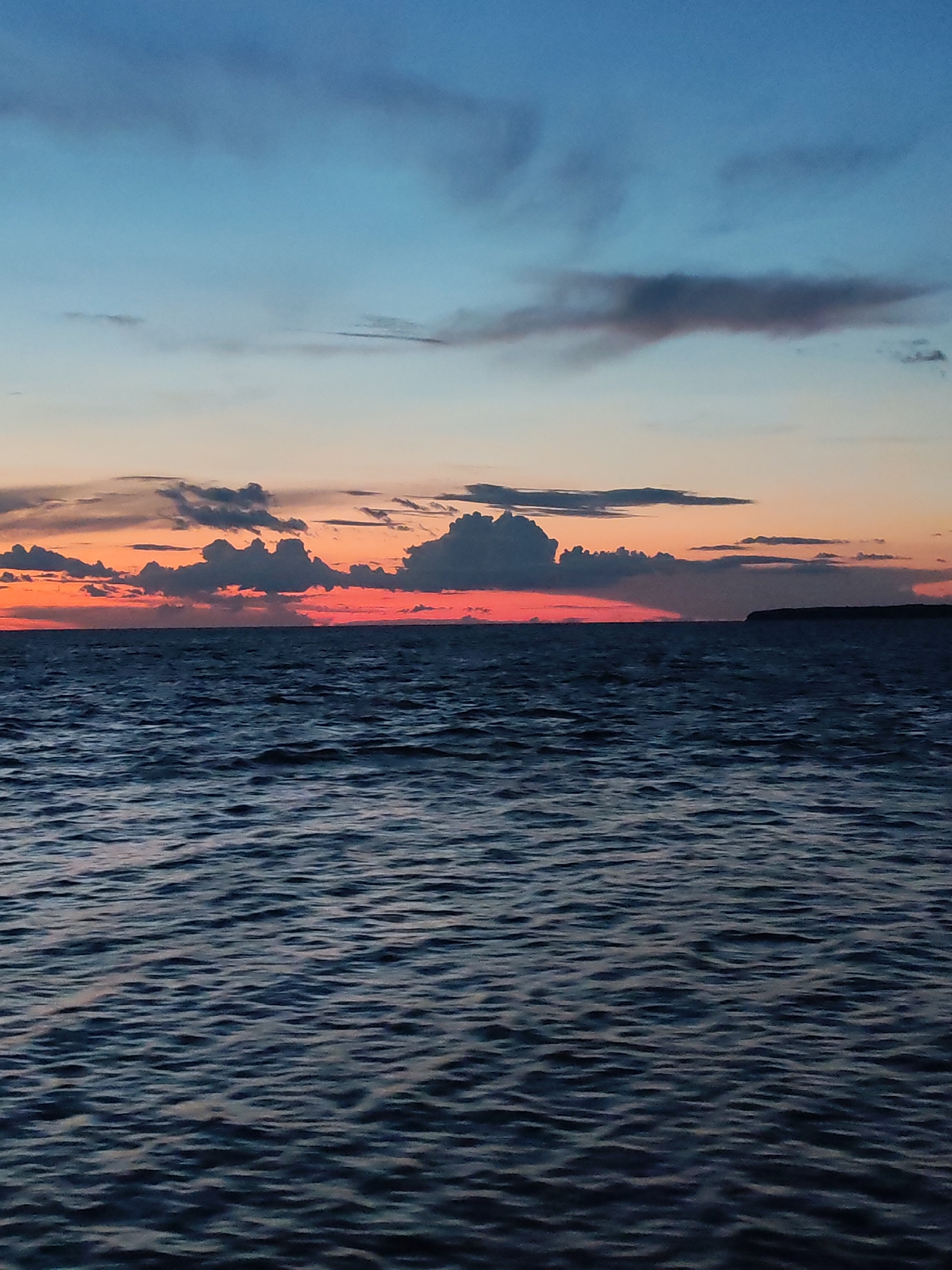 The image depicts a serene sunset over a body of water. The sky transitions from a deep blue at the top to vibrant hues of orange and pink near the horizon, with scattered dark clouds. The water is dark blue and exhibits gentle ripples. In the distance, a dimly lit landmass or island is visible on the horizon. The scene appears to be a natural landscape, with no urban elements or people present. The location of Pochinok, Russia is not discernible from the visual information provided in the image.