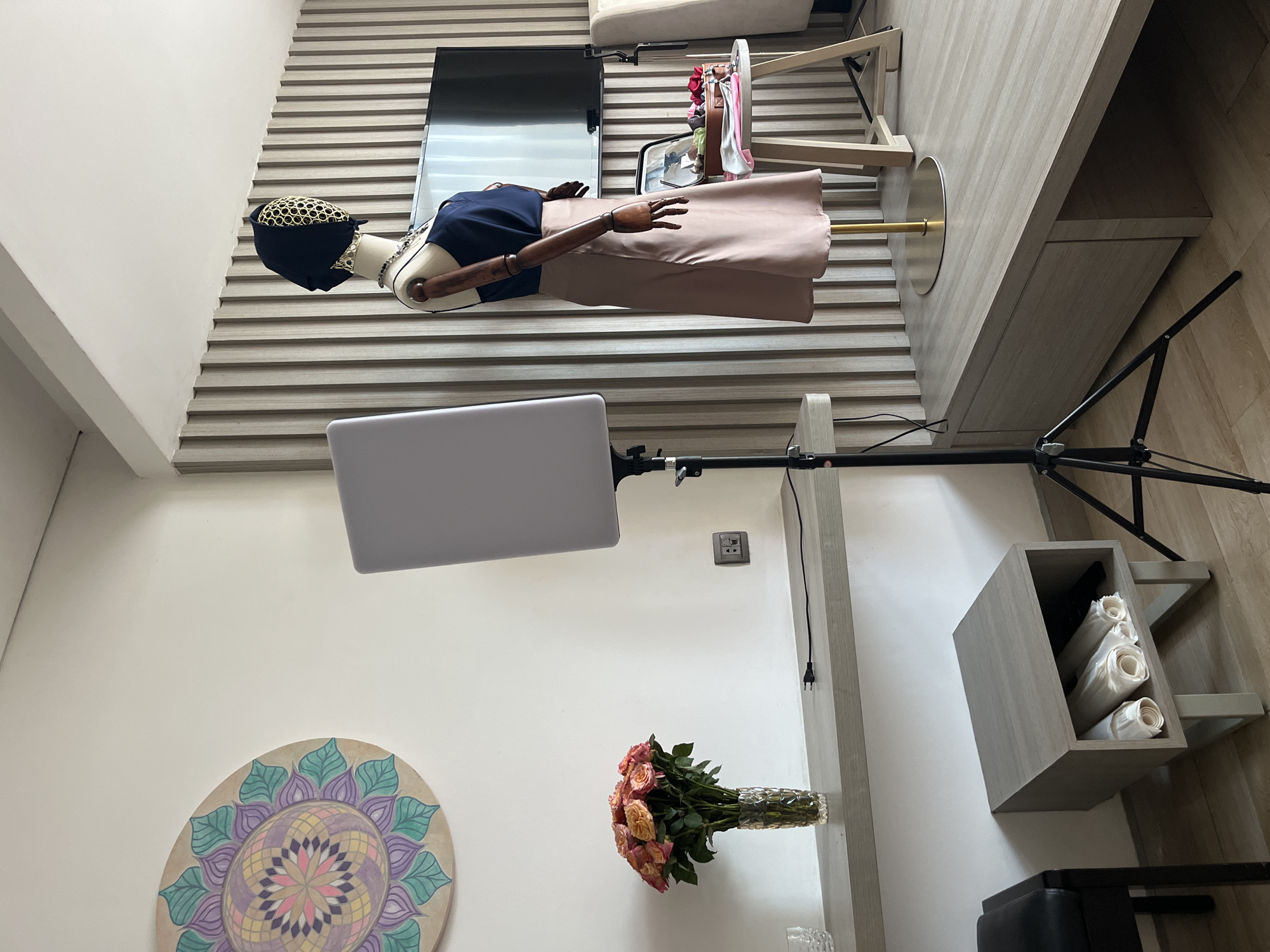 The image appears to be taken inside a retail or studio space in Bac Nha Trang Ward, Vietnam. A female mannequin wearing a dark blue top, beige skirt, and dark head covering with gold trim is the primary subject. The mannequin is posed against a wall with horizontal beige slats, and a television is mounted behind it.
A lighting setup with a rectangular light panel on a tripod is positioned to illuminate the mannequin. A round mandala-style artwork hangs on the wall. A vase of peach-colored roses is also present on the wall.
A clothing rack with a few items is located near the mannequin. A wooden cabinet with folded material is visible on the floor, and a chair with black padding is also in view. The floor is a light-colored wood.