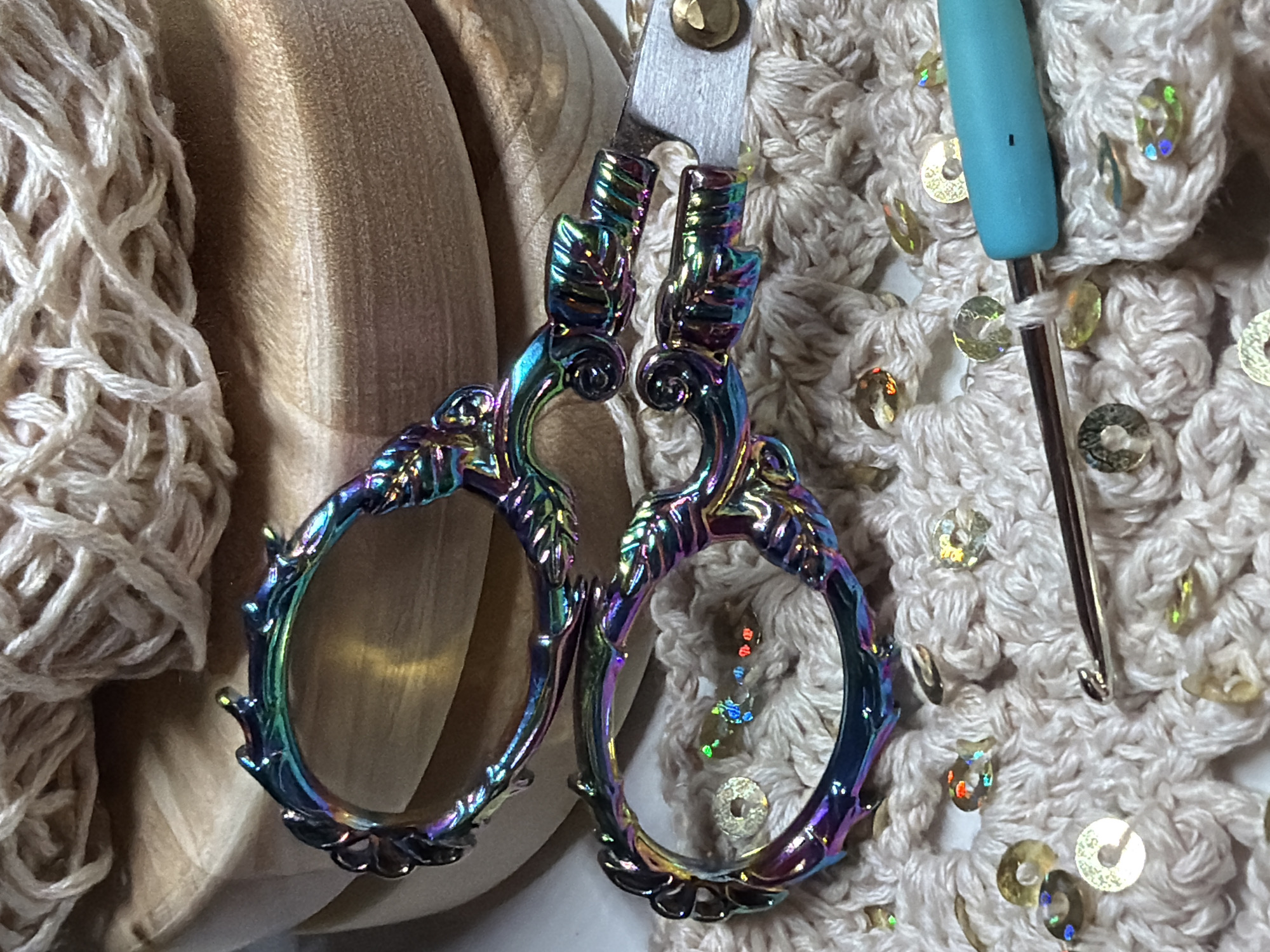 The image shows a close-up of crafting materials. From left to right, there is a ball of cream-colored yarn, a portion of a velvet-covered craft pumpkin, iridescent scissors hanging from a silver-colored metal loop with a gold rivet, a crocheted piece with sewn-on sequins, and a light blue-handled crochet hook. The scene appears to be indoors. There is no visual indication of the location being in Chaikovsky, Russia.