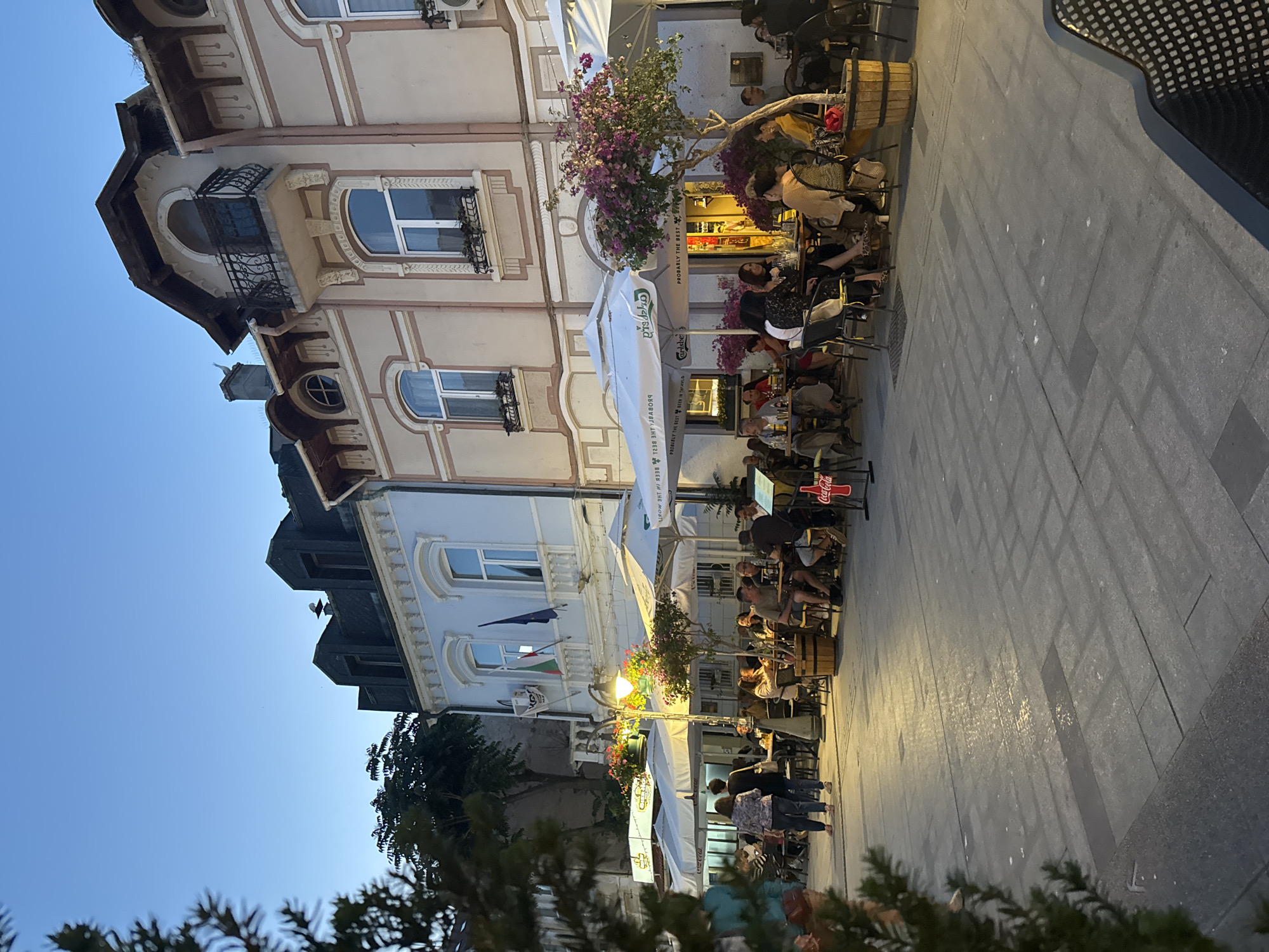 The image depicts an outdoor dining area in Burgas, Bulgaria, likely on a pedestrian street. The scene is illuminated by streetlights and the ambient glow of the late evening sky. Several tables and chairs are set up under white umbrellas, with diners seated and engaged in conversation and eating. Buildings with architectural details line the street, displaying a mix of traditional and modern elements. A Coca-Cola advertisement sign is visible. The pavement is made of stone tiles, and trees and potted plants add to the ambiance. The overall impression is one of a lively and pleasant evening in a European city.