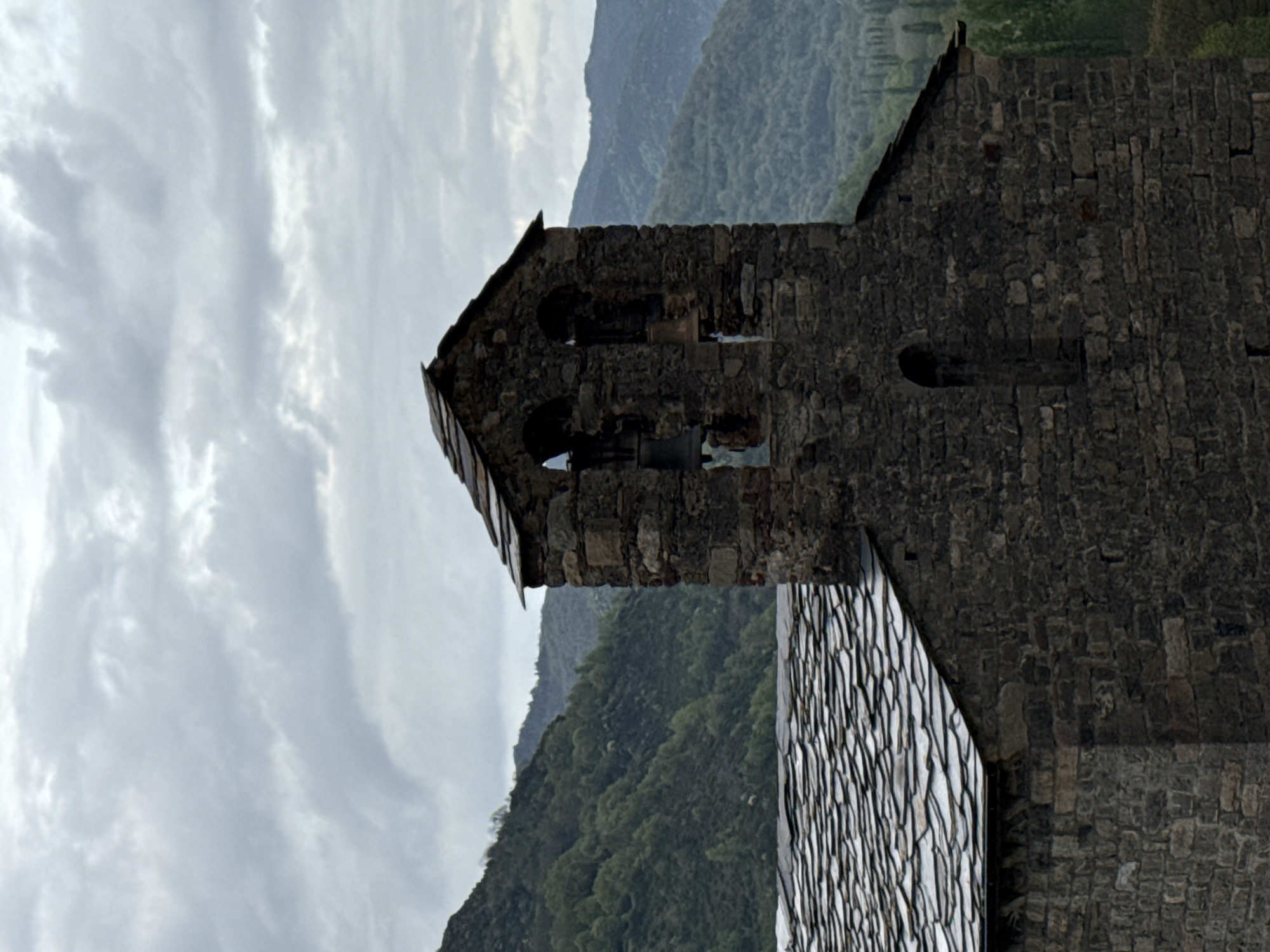 An exterior shot of the Romanesque Church of Sant Romà de Vila in Villanova, Spain, showing a stone church building with a stone tile roof. The sky is cloudy and mountains are visible in the background.