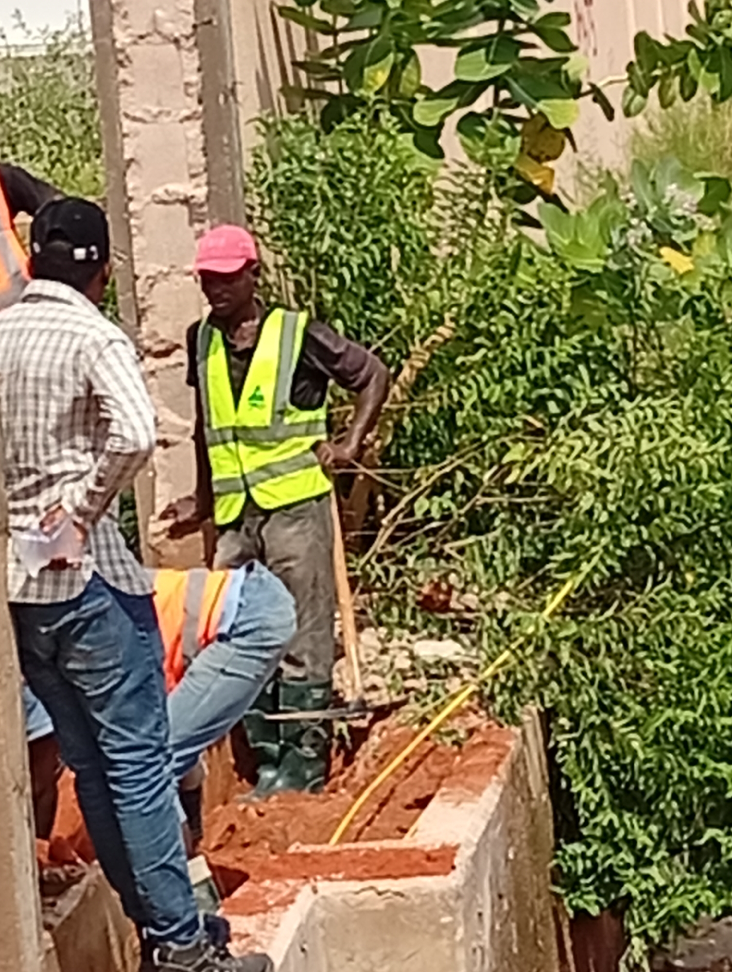 In Gombe, Nigeria, a construction scene is depicted with several individuals engaged in work. One man, wearing a pink cap and a bright yellow safety vest over a dark shirt, stands near a partially constructed brick structure. He is holding a wooden tool, possibly a shovel or tamping rod, and appears to be working at ground level. Another person, to the left, is dressed in a plaid shirt and blue jeans, facing the man in the safety vest. Their stance suggests interaction or observation of the ongoing work. Further to the left, another individual, partially visible, wears a black cap and a plaid shirt, with their back to the camera. In the foreground, a segment of the construction project reveals exposed red bricks and a trench. A yellow cable or pipe is visible running across the scene, implying utility work. The background is filled with lush green vegetation and a textured wall, suggesting an outdoor environment in a developing area.