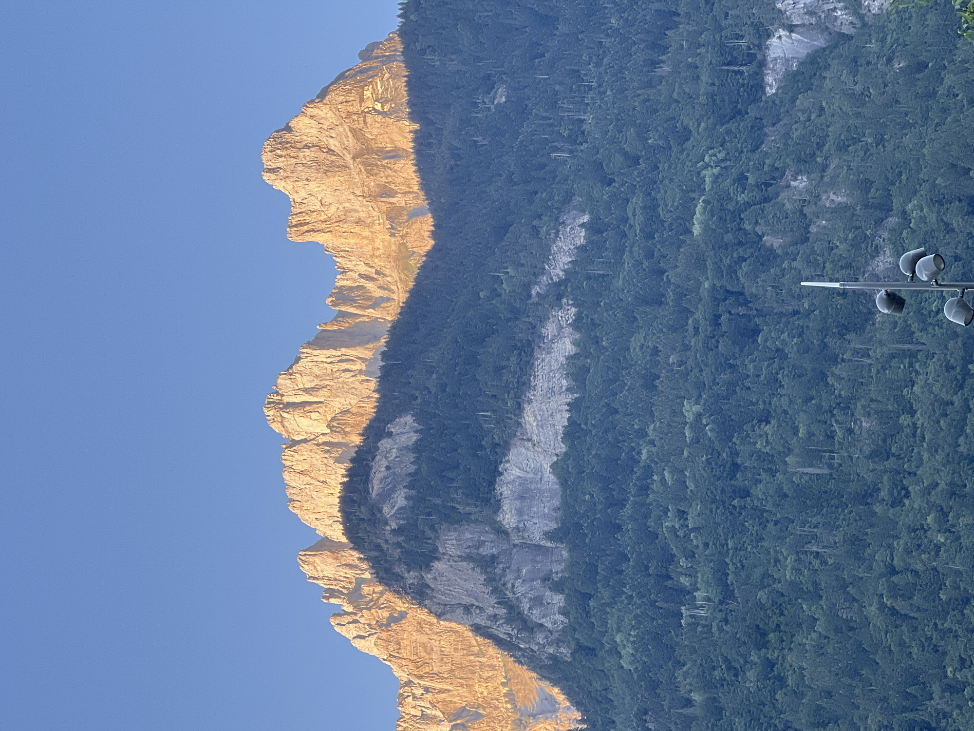 The image shows a majestic mountain range, likely in Sallanches, France, with a clear blue sky. The sun is low in the sky, casting a warm, golden light on the rugged, rocky peaks of the mountain's western face. The eastern face of the mountain is heavily forested with dark green trees, creating a stark contrast between the illuminated rock and the shaded forest. The lighting suggests it is either early morning or late afternoon. The mountain itself appears to be a significant natural landmark, characterized by its dramatic cliffs and dense forestation. Towards the bottom right of the image, a modern street lamp with multiple lights is visible, hinting at the presence of human infrastructure or a viewpoint area near the mountain. No people are visible, and there is no text in the image. The overall mood is one of natural grandeur and peacefulness.