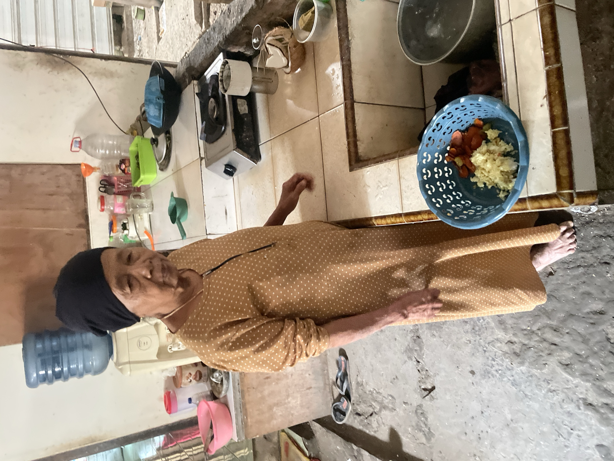 In a modest kitchen in Parakan, Indonesia, an elderly woman with a black headscarf and a polka-dot dress stands by a counter. To her left, a blue water dispenser sits atop a white appliance. The counter is tiled in beige and brown, with various kitchen items scattered about, including a gas stove, bowls, and containers. A blue colander filled with chopped vegetables sits near the sink. The floor is concrete, showing signs of wear and tear. The overall impression is one of a lived-in, functional kitchen.