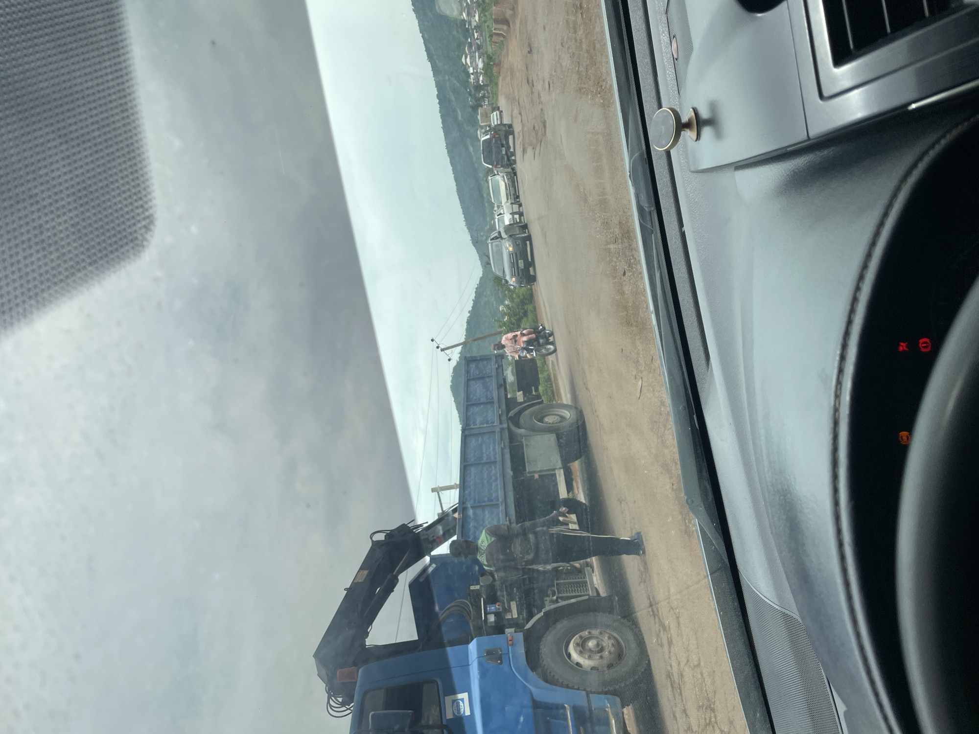 The image shows a street scene in Koton Karifi, Nigeria. The perspective is from inside a vehicle, looking out through the windshield. The day appears overcast, with a dull, diffused light suggesting it might be late morning or early afternoon, or perhaps a cloudy day.
The main focus of the image is a large blue truck with a crane attached. Two people are visible near the truck. One person, wearing a light-colored shirt and dark pants, is standing on the ground near the rear of the truck, appearing to be directing or observing. Another person, wearing a green reflective vest and dark clothing, is positioned on the truck itself, near the crane. This suggests some kind of operation is taking place involving the truck and its equipment, possibly loading or unloading.
In the background, a line of vehicles is parked along a dirt road. These appear to be mostly SUVs and cars. Beyond the road, a hillside is visible with scattered buildings and vegetation, indicating a semi-urban or rural setting. A motorcycle with a rider is also present on the road, adding to the sense of everyday activity.
The interior of the vehicle from which the photo is taken is partially visible, including a dashboard with some controls and a circular attachment on the dashboard. The overall scene depicts a moment of work or traffic on a road in Nigeria. There is no discernible text in the image.