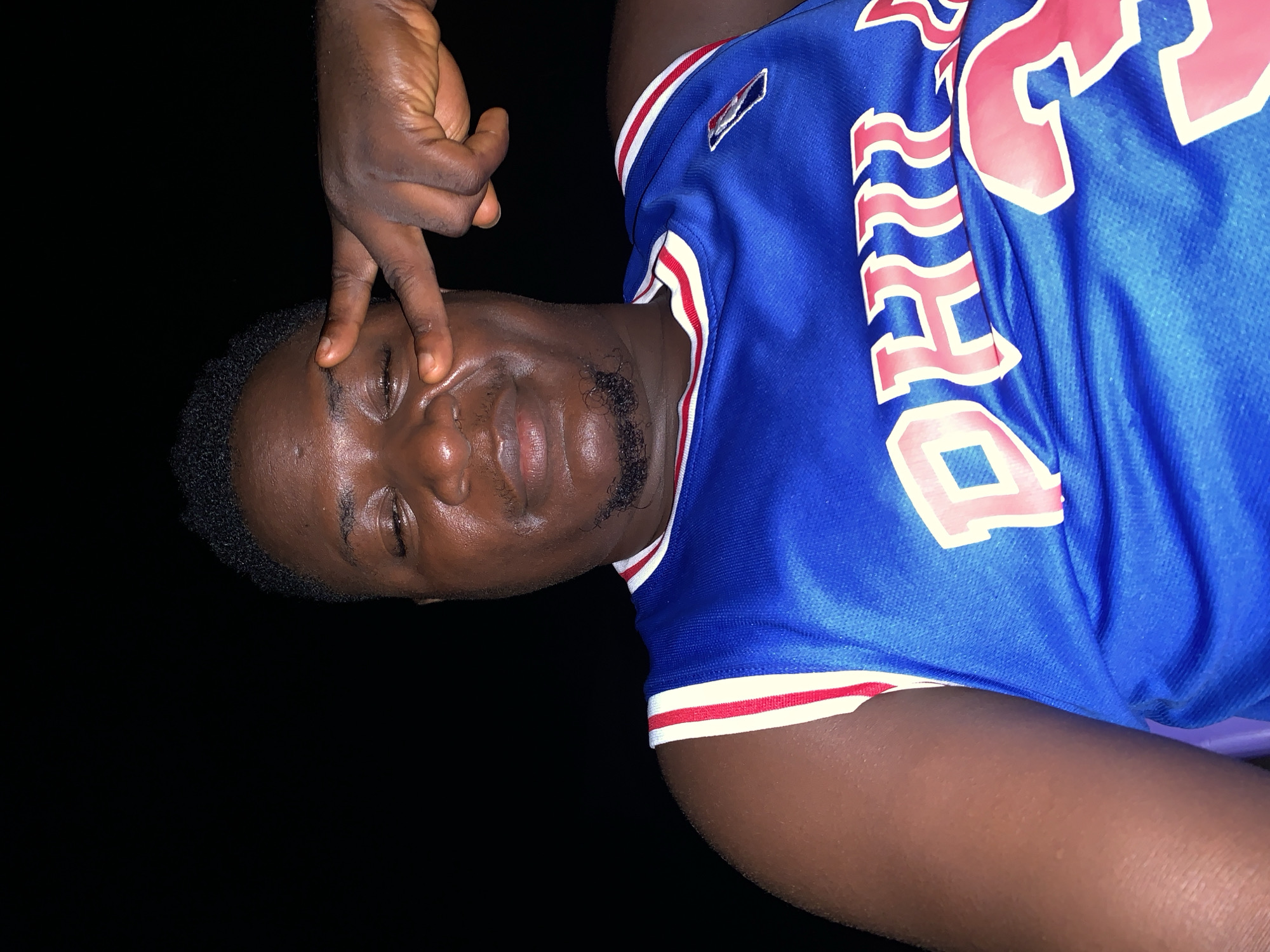 A person with dark skin and short, curly black hair is depicted in a medium close-up shot against a stark black background. The image is rotated 90 degrees clockwise. The subject is wearing a blue sports jersey with red and white striped trim on the sleeves and collar. Partially visible text on the jersey, in red with a white outline, reads "PHILA" and a number, possibly '23' or '33'. A small red, white, and blue emblem is also present on the upper right shoulder area of the jersey.
The person has a trimmed goatee or beard. Their right hand is raised, with the index and middle fingers extended and pressed against their forehead, near the right eyebrow. The eyes are partially closed or squinting, and the facial expression is neutral. A slight sheen is visible on the skin, suggesting direct lighting like a flash, and possibly sweat or oil. The black background indicates a dark environment, likely night or a very dimly lit indoor setting in Akwanga, Nigeria. The hand gesture is a prominent feature of the pose.