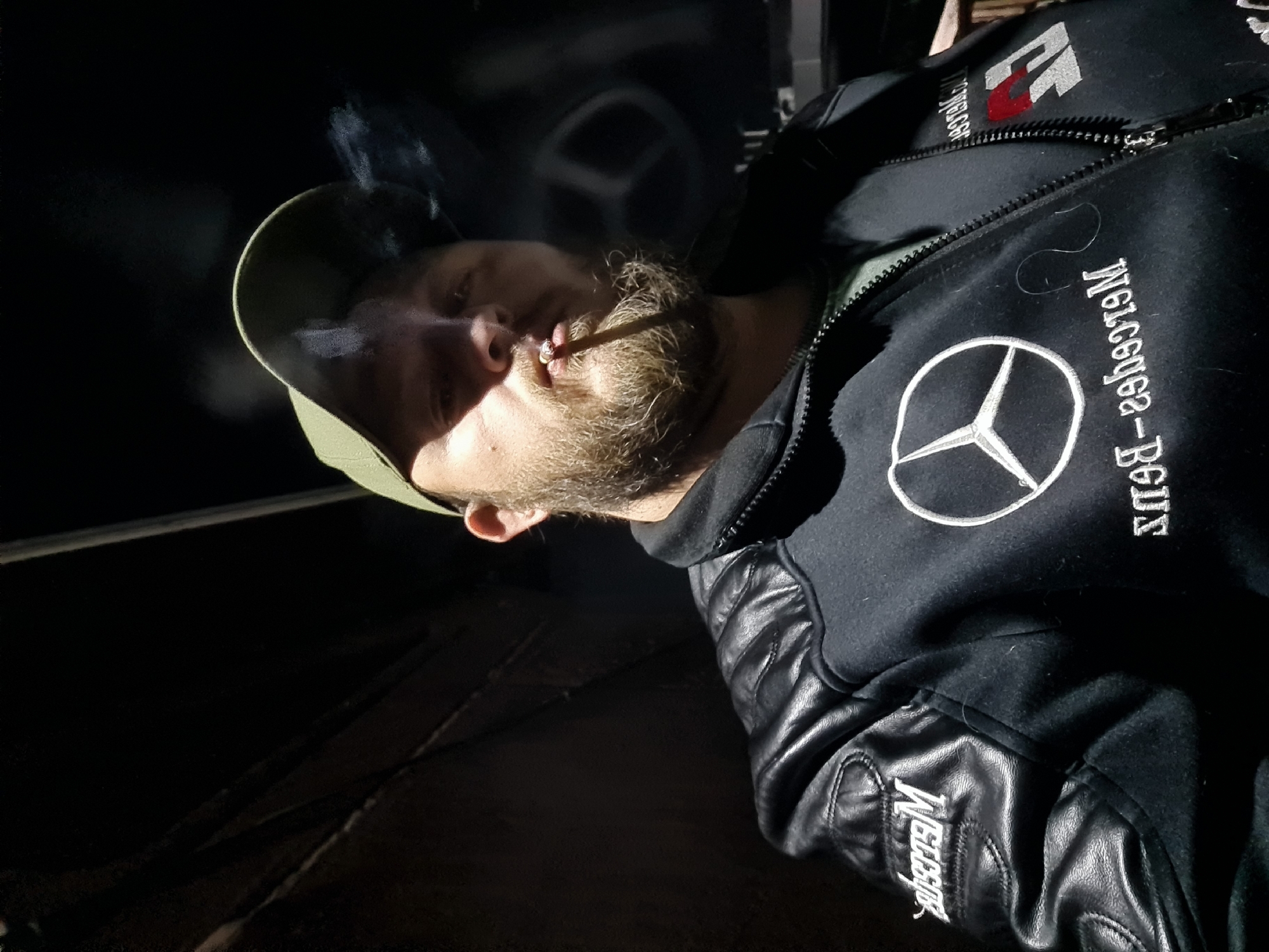 A bearded man in a green baseball cap and a black jacket with the Mercedes-Benz logo is smoking. The background is dark, with an indistinct circular shape behind him.
