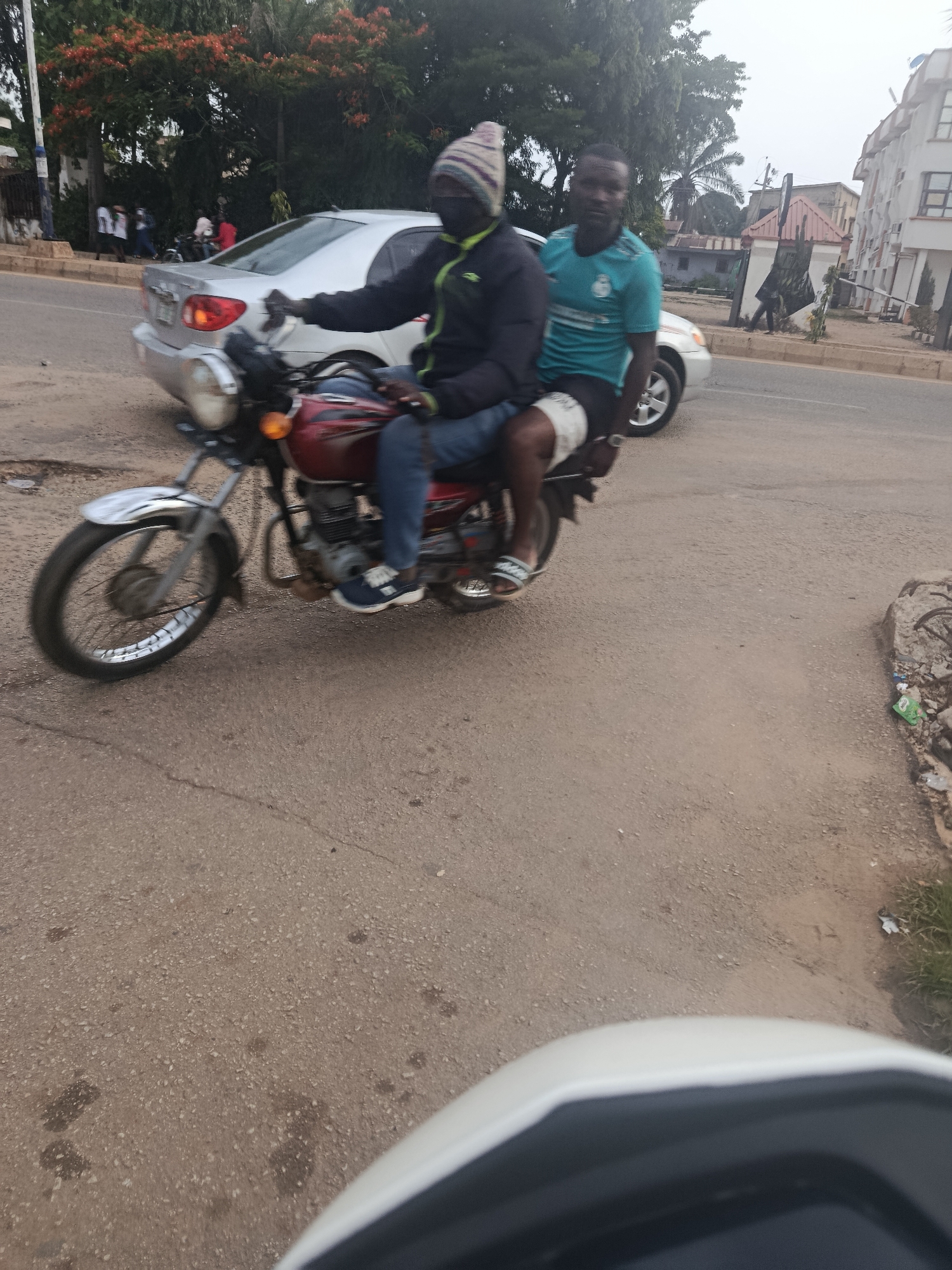 Two men ride a red motorcycle on a paved road in New Stadium, Nigeria. The driver wears a dark jacket with green accents and a knitted hat. The passenger, wearing a blue t-shirt, sits behind him. A silver car is visible in the background, along with trees and buildings. The road appears to be in moderate condition, with some cracks and debris. The lighting suggests it is daytime.