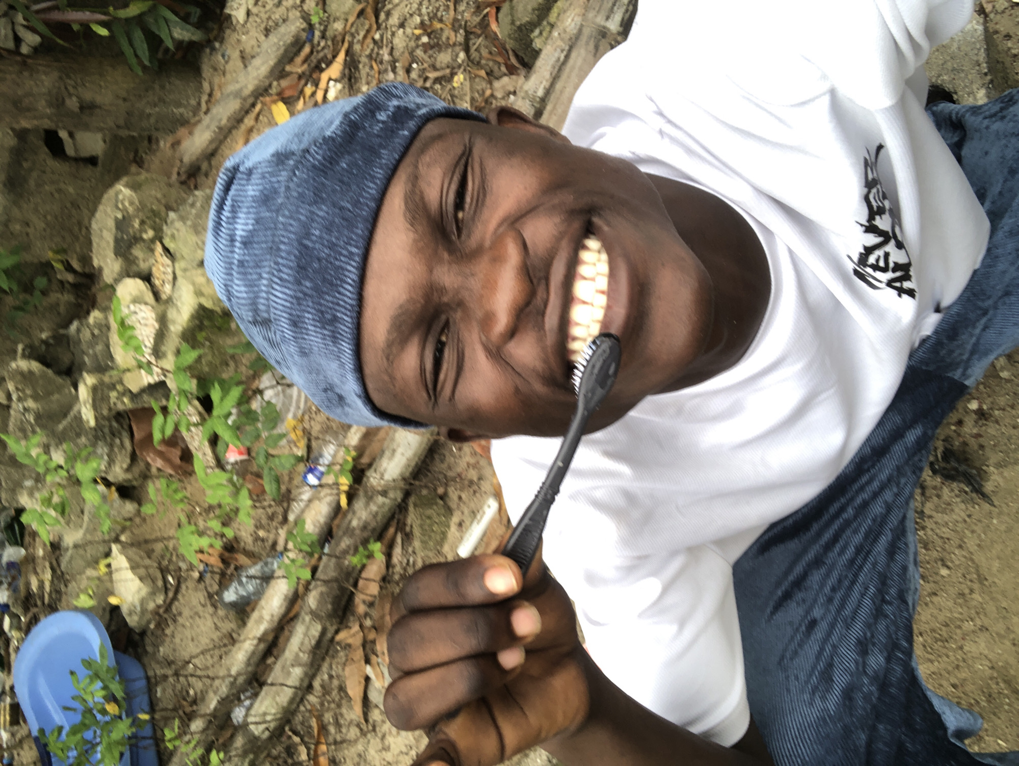A young Black male wearing a blue beanie, a white t-shirt, and blue jeans is shown from the chest up, holding a black toothbrush to his teeth. He is smiling broadly, exposing his teeth. The background appears to be an outdoor, somewhat disheveled area with rocks, dirt, fallen leaves, and some green foliage. A blue plastic basin is visible in the upper left corner. The overall scene suggests a casual, informal setting, possibly in Ado, Nigeria, given the context provided.