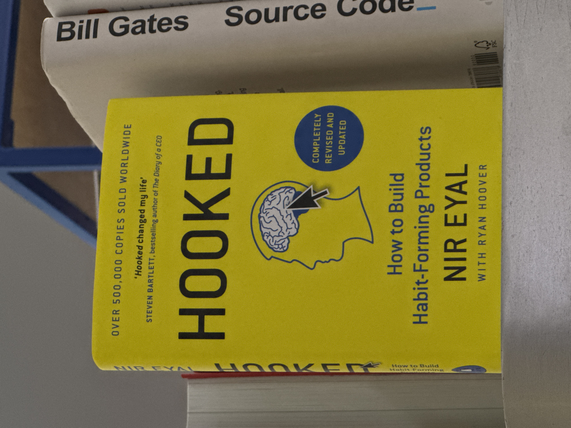 The image shows a close-up of two books stacked on top of each other. The top book is titled "Bill Gates Source Code." The second book is yellow and titled "HOOKED" in large, bold black letters. The book cover also features a stylized illustration of a human head with a brain inside, and the words "How to Build Habit-Forming Products" and "NIR EYAL WITH RYAN HOOVER." Additional text on the cover indicates "OVER 500,000 COPIES SOLD WORLDWIDE" and "Completely Revised and Updated." The location and specific details about Yerevan, Armenia are not evident in the image.
