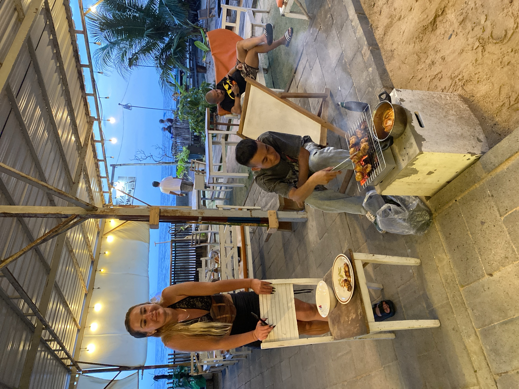 A scene unfolds outdoors under a covered patio near the ocean in Mũi Né, Vietnam. In the foreground, a woman with blonde hair stands beside a white wooden chair, leaning on its back. She is wearing a black lace top and a black skirt. On the seat of the chair, a white bowl and a plate with food are placed. To the right of the woman, a man with short dark hair is grilling skewers of meat over a small charcoal grill. He is wearing a grey t-shirt and light-colored pants. Next to the grill, a metal pot is situated on a concrete block. Further back, a man with a shaved head is reclining on an orange lounge chair, wearing a black t-shirt and patterned shorts, and looking at his phone. The environment includes tables and chairs set up on a tiled floor, with the ocean and a wooden fence visible in the background. Palm trees are also present, suggesting a tropical setting. The sky is a dusky blue, indicating it is either early morning or late evening.