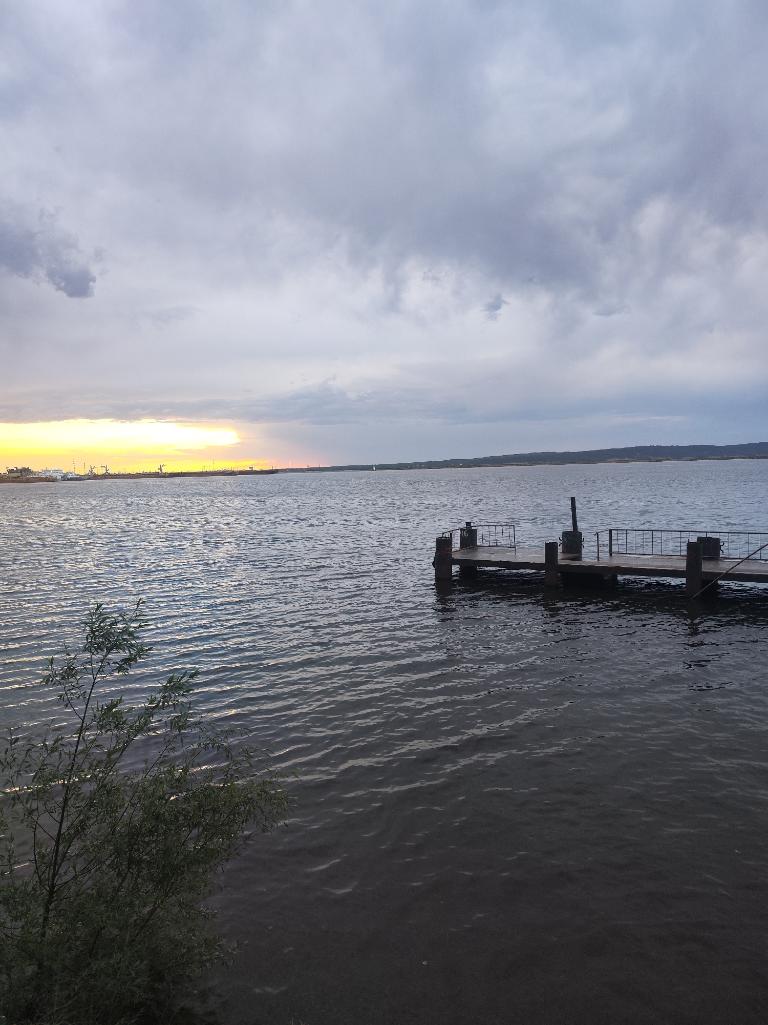 This image depicts a body of water, likely a lake or river, with a small wooden pier extending into it. The sky is overcast with grey clouds, but there is a sliver of golden light visible on the horizon, indicating either sunrise or sunset. In the distance, there is a suggestion of an industrial area, possibly a port or factory. In the foreground, a tree or bush is visible on the bank. The photograph appears to be taken from the city of Chaikovsky, Russia, based on the industrial structures and water body location. The water is calm with slight ripples. There are no people visible in the frame.