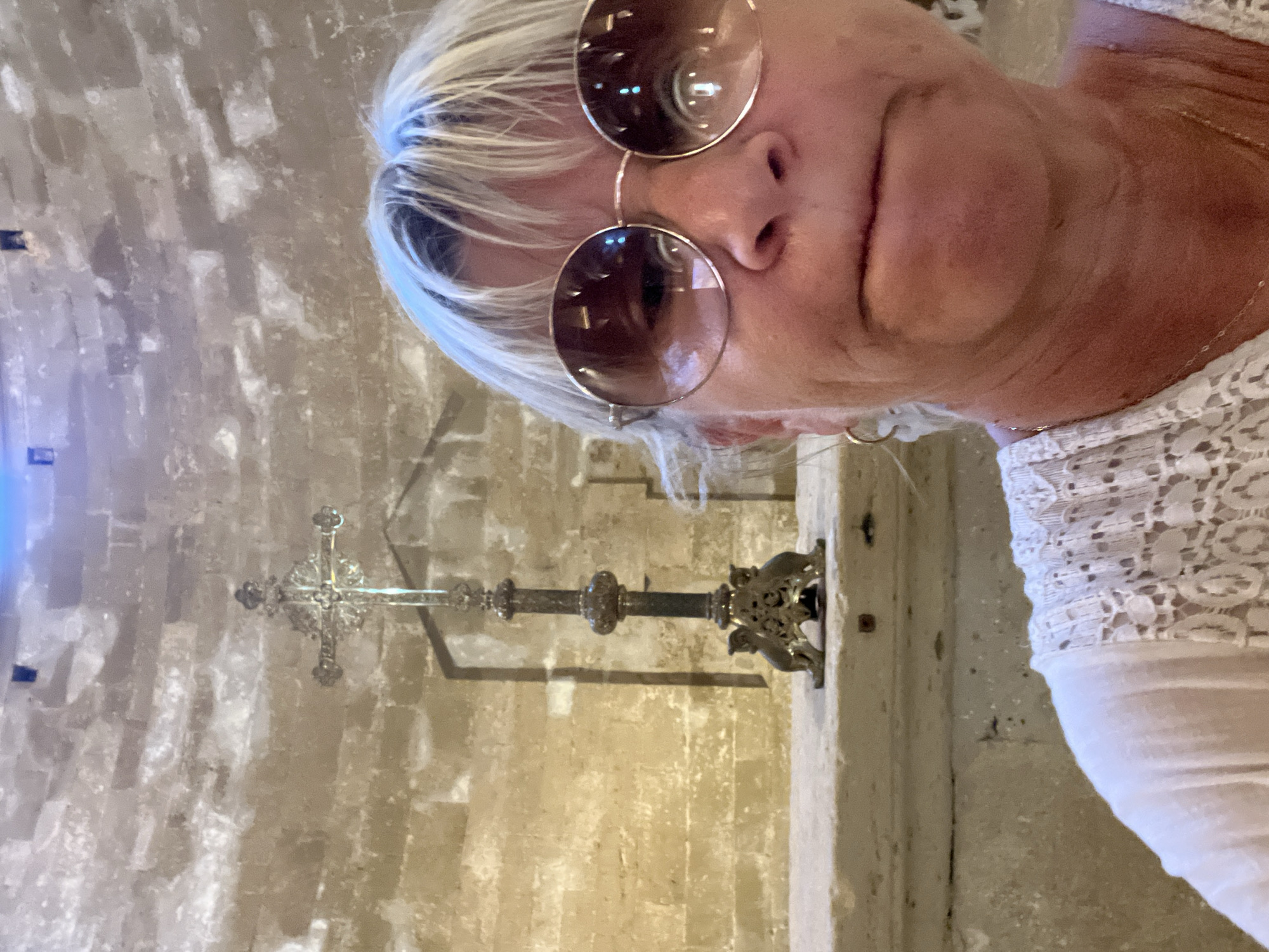 The image shows a medium shot of a woman taking a selfie in what appears to be the interior of a historic church. The woman, with blonde hair and wearing large, round sunglasses and a white lace top, is positioned in the foreground, facing the camera with a slightly pursed expression. Behind her, a stone wall forms the background, and a decorative, ornate metal cross on a stand is visible on a stone ledge. The architectural style and stonework suggest an old religious building. The lighting is somewhat dim, with a blueish hue visible at the top of the frame, possibly from artificial lighting or stained glass. The woman's attire and the sunglasses indicate she is likely a tourist. The scene is likely within the Cathedral Saint-Pierre de Maguelone, located on an island off the coast of Villeneuve-lès-Maguelone, France, given its historical significance and architectural features.