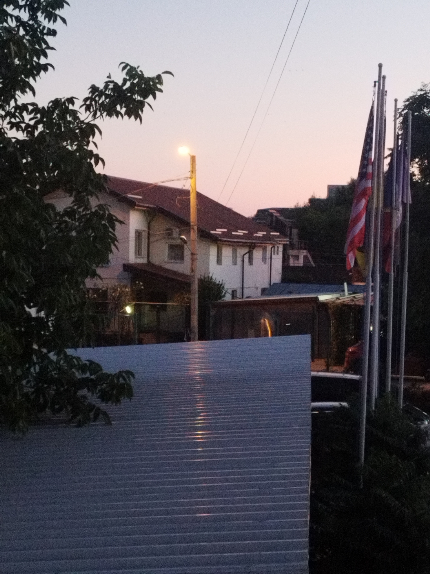 The image captures a twilight scene in Bucharest, Romania, featuring a residential building with a red-tiled roof, a streetlamp, and several flags on flagpoles. The American flag is prominently displayed among other flags, suggesting a diplomatic or official setting. A dark-leaved tree with branches partially obscures the view of the house. In the foreground, a corrugated metal roof extends across the bottom of the frame, reflecting some light. The sky is a gradient of pale purple and orange, typical of dusk. A few thin electrical wires are visible against the sky. The overall impression is one of a quiet, possibly residential or institutional, area at the end of the day. There are no visible people or animals in the frame.