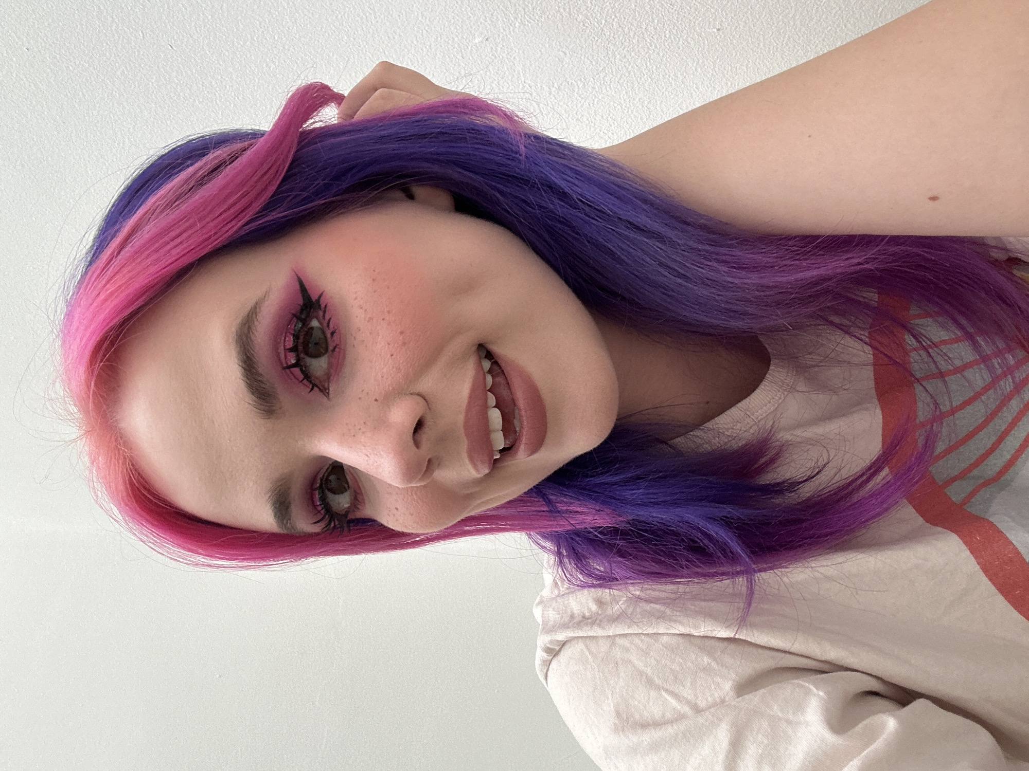 A fair-skinned individual with pink and purple hair, wearing pink eye makeup and lipstick, poses indoors. The individual has freckles on their face and is smiling. They are wearing a light-colored t-shirt with a faded red design. The background is a plain white wall. The location in the city of City of Zagreb, Croatia is not discernible from the image.