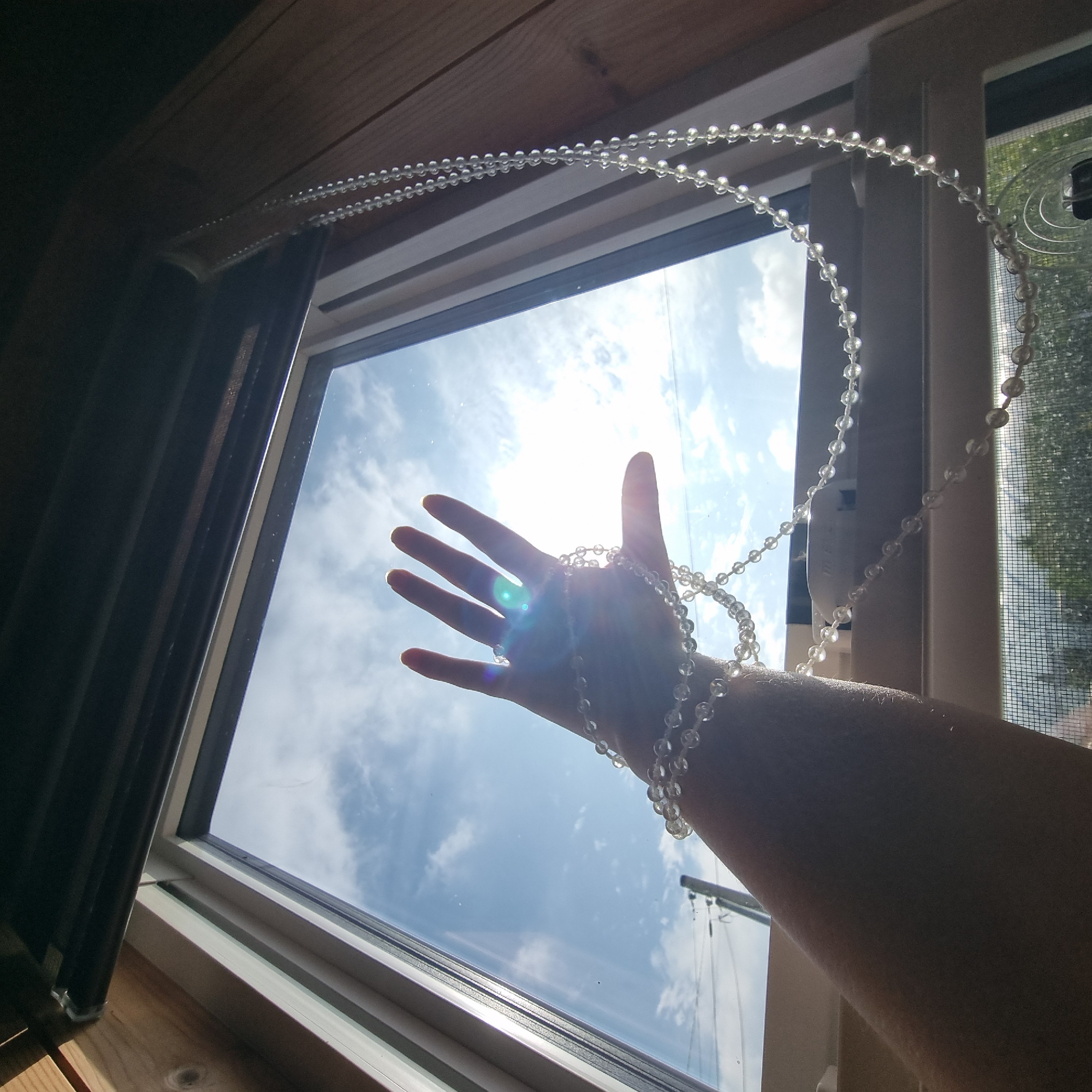 The image shows a human hand reaching towards a sunlit window. A string of clear beads is draped around the wrist and hand, extending downwards and out of frame. The hand is silhouetted against a bright blue sky with scattered clouds, and the sun is creating a lens flare effect. The window appears to be in a room with wooden walls and a dark-colored roller blind. In the background, through the window, faint outlines of utility poles and trees are visible. The overall scene suggests an indoor setting looking out at a bright day. There is no indication of the location being Dunnae-myeon, South Korea.