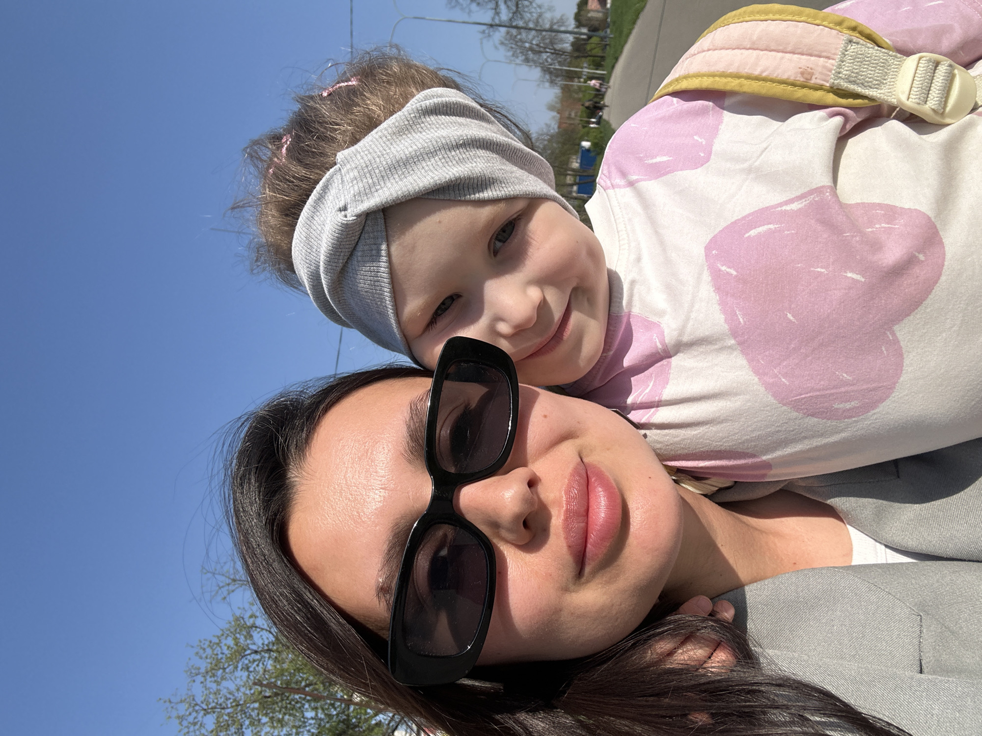 The image shows a selfie of a woman and a child outdoors. The woman is wearing black sunglasses and a grey jacket, and her dark hair is visible at the bottom of the frame. The child is being carried on the woman's back, wearing a grey headband, a light-colored shirt with pink heart patterns, and a backpack-style carrier. The sky is bright blue and sunny. The background suggests an urban park or roadside environment with trees and a paved area. It is likely the location is Chaikovsky, Russia.