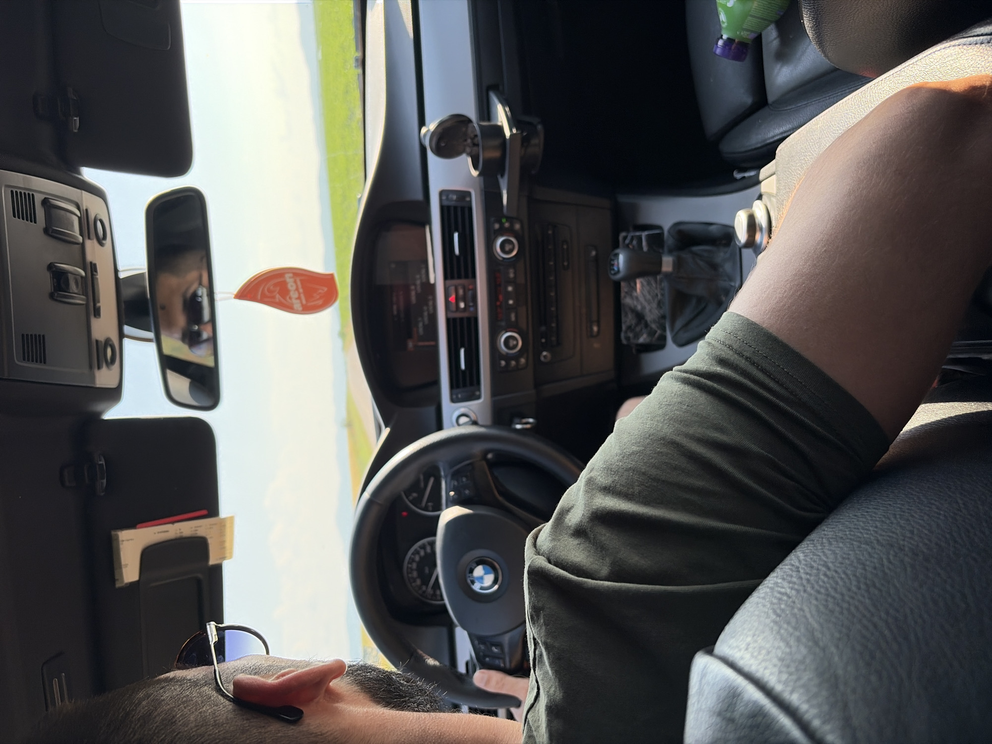 The image depicts the interior of a car from a rear-passenger perspective, looking forward. A male driver, with dark hair, a beard, and sunglasses, is visible from behind, wearing a dark green t-shirt, with his hands on the steering wheel. His reflection, also wearing sunglasses, is visible in the rearview mirror. The vehicle is identified as a BMW by the steering wheel emblem and dashboard design, featuring a manual transmission, an infotainment screen, and climate controls. A red "areon" brand air freshener hangs from the rearview mirror. A paper document is clipped to the driver's sun visor. An accessory cup holder/ashtray is attached to the dashboard on the passenger side, where a green bottle is also visible on the seat. The car is on a paved road, driving through a rural landscape characterized by bright green fields and distant hills under a clear blue sky. The scene is located in the city of Lyubimets, Bulgaria.