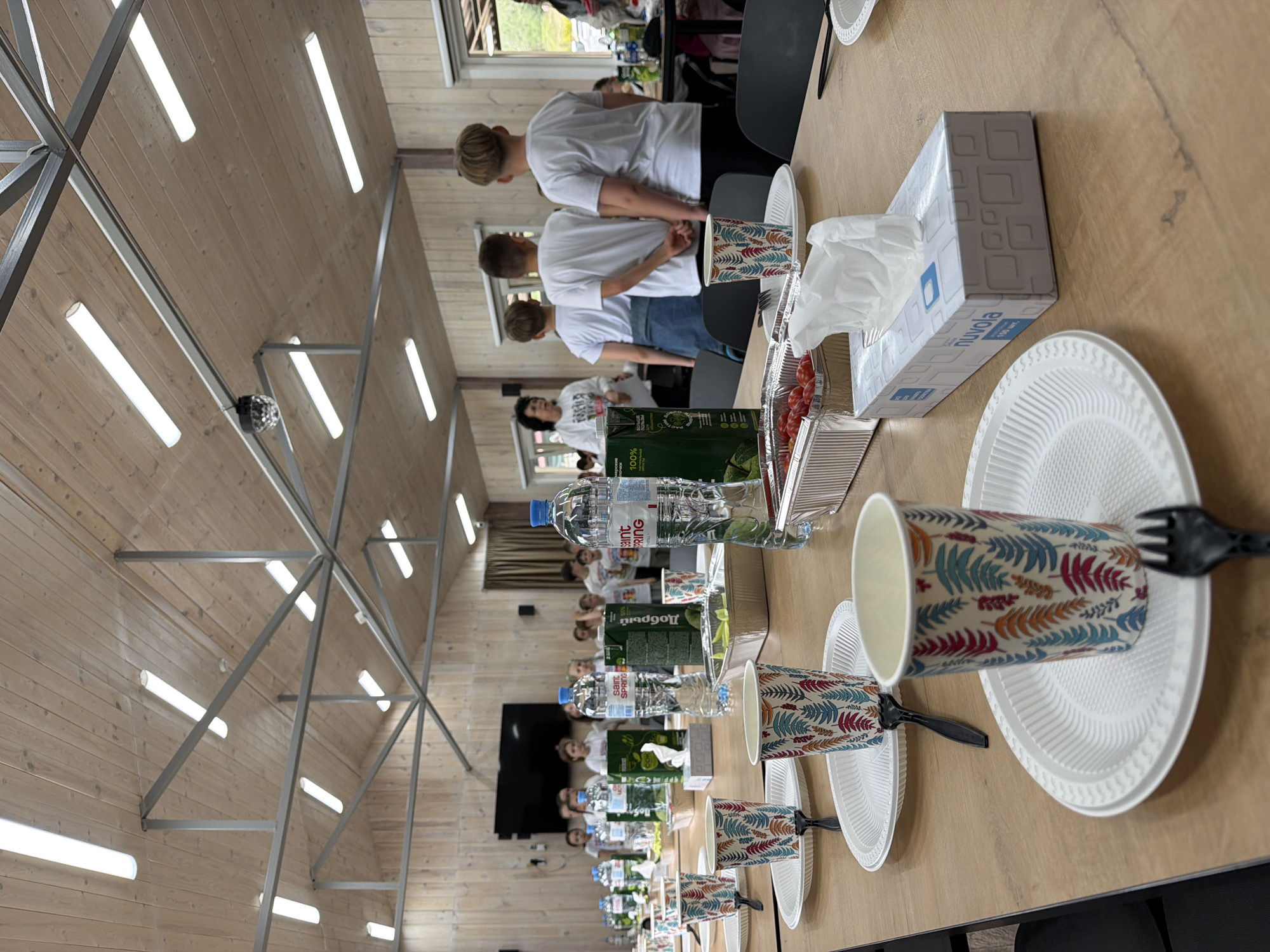 The image shows a brightly lit, indoor space, possibly a dining hall or conference room. A long wooden table stretches through the center of the frame, set with a row of disposable white plates, patterned paper cups, plastic forks, water bottles, juice boxes, and a tissue box.
Several people, mostly wearing white shirts, are standing along the side of the table, facing away from the camera. The space has a wooden ceiling with a network of metal beams and fluorescent lights. A black TV screen and window is visible in the background. The location is not evident from the image.
