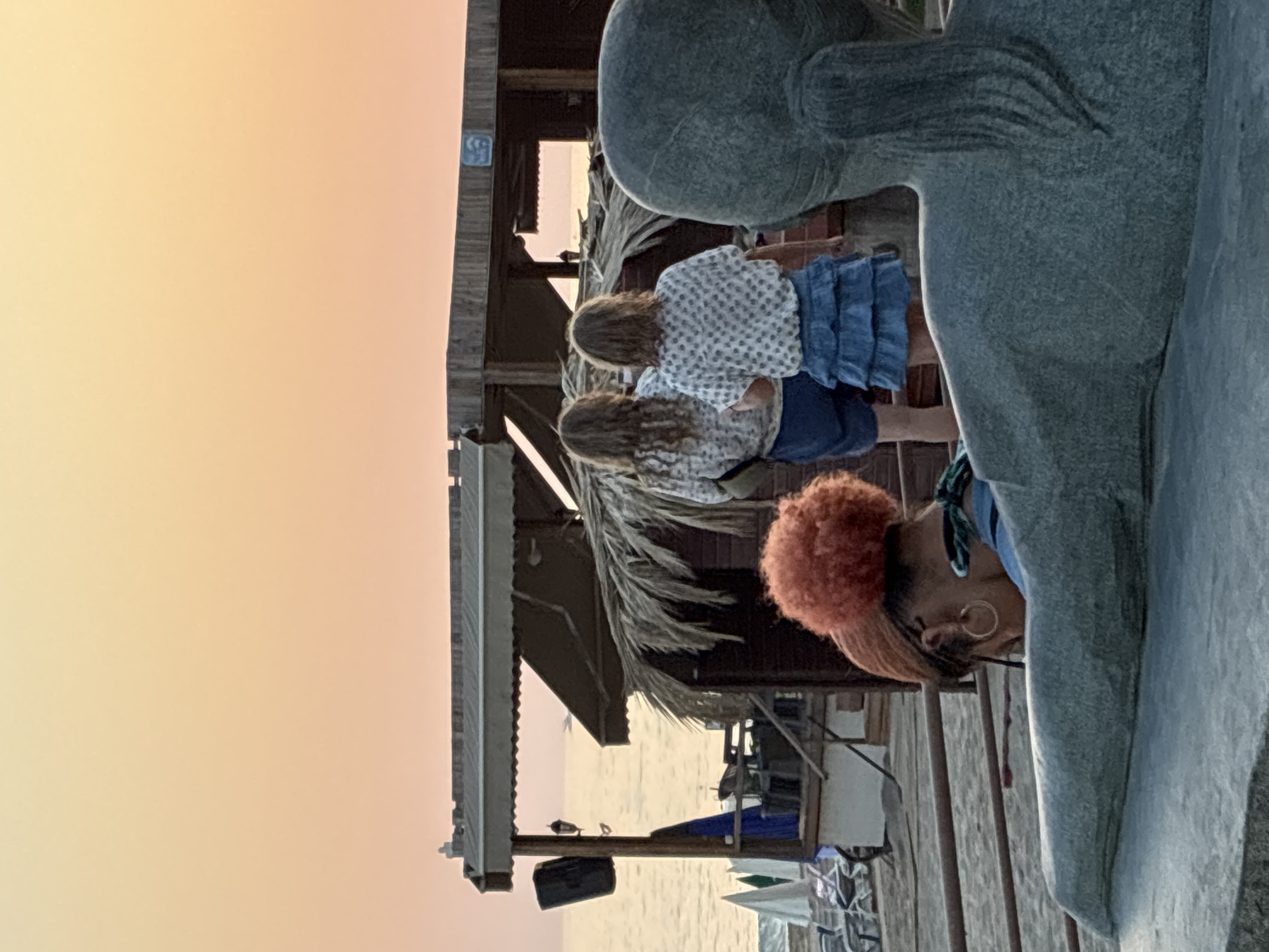The image shows a scene at dusk, with a soft, peachy sky. In the foreground, a large, grey, statue-like sculpture dominates the right side of the frame, with its texture suggesting it's made of stone or concrete. To the left of the sculpture, a person with bright orange, curly hair and hoop earrings is visible from the side, wearing a grey hoodie and a blue scarf around their neck. They are looking down, their face partially obscured.
Behind the sculpture and the person, there is a wooden structure resembling a beach hut or pier, with a thatched roof. Two young girls, seen from the back, are standing near this structure. They both have long, wavy, light brown hair and are wearing similar white tops with a small dark pattern and blue denim skirts. They appear to be looking out towards a body of water, possibly the sea.
Further in the background, a faint silhouette of a person can be seen on what looks like a beach. The overall setting seems to be a waterfront area, possibly a resort or a public beach. The time of day is clearly indicated by the warm, diffused light of the sunset or sunrise. There are no discernible emotions from the subjects, as their faces are either not visible or turned away. No visible text is present in the image. The scene evokes a peaceful, end-of-day atmosphere.