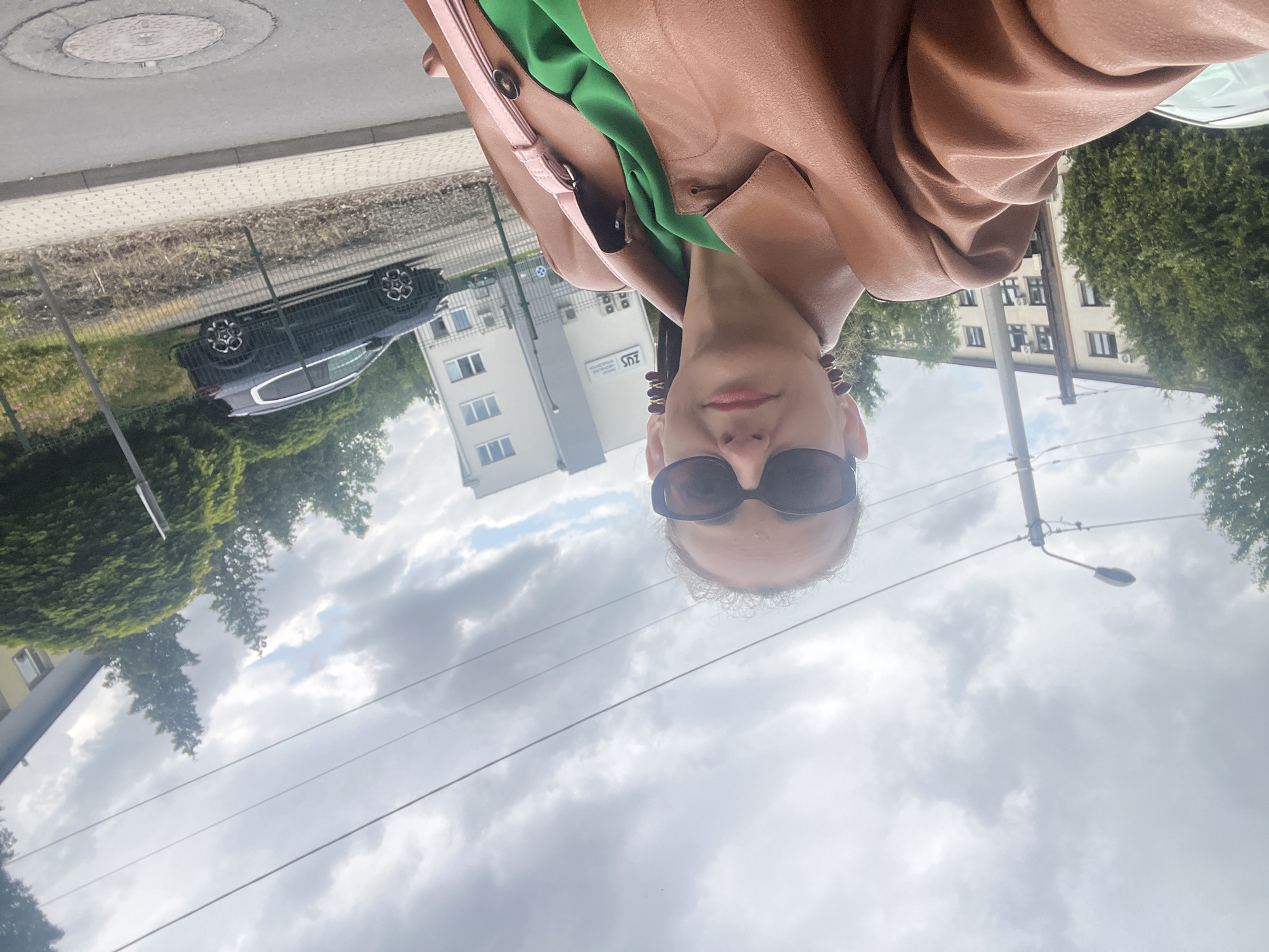 A medium shot photograph captures a woman from the chest up, standing outdoors under a cloudy sky. She is wearing oversized dark sunglasses, a brown leather jacket with exaggerated shoulder pads, and a vibrant green top. Her hair is pulled back, and she has distinctive, stacked brown earrings. A pink strap from a bag is visible across her chest.
The background suggests an urban or suburban setting. To the left, a light-colored building with multiple windows and visible air conditioning units is partially obscured by lush green trees. Further back, a taller, light-colored building with a flat roof and more windows is visible. A street lamp with multiple lights stands to the left of the woman, with power lines running across the sky. A dark-colored SUV is parked on the right side of the image, behind a metal fence. The ground in the foreground is paved with light gray interlocking pavers, and a circular manhole cover is visible in the lower right corner.
