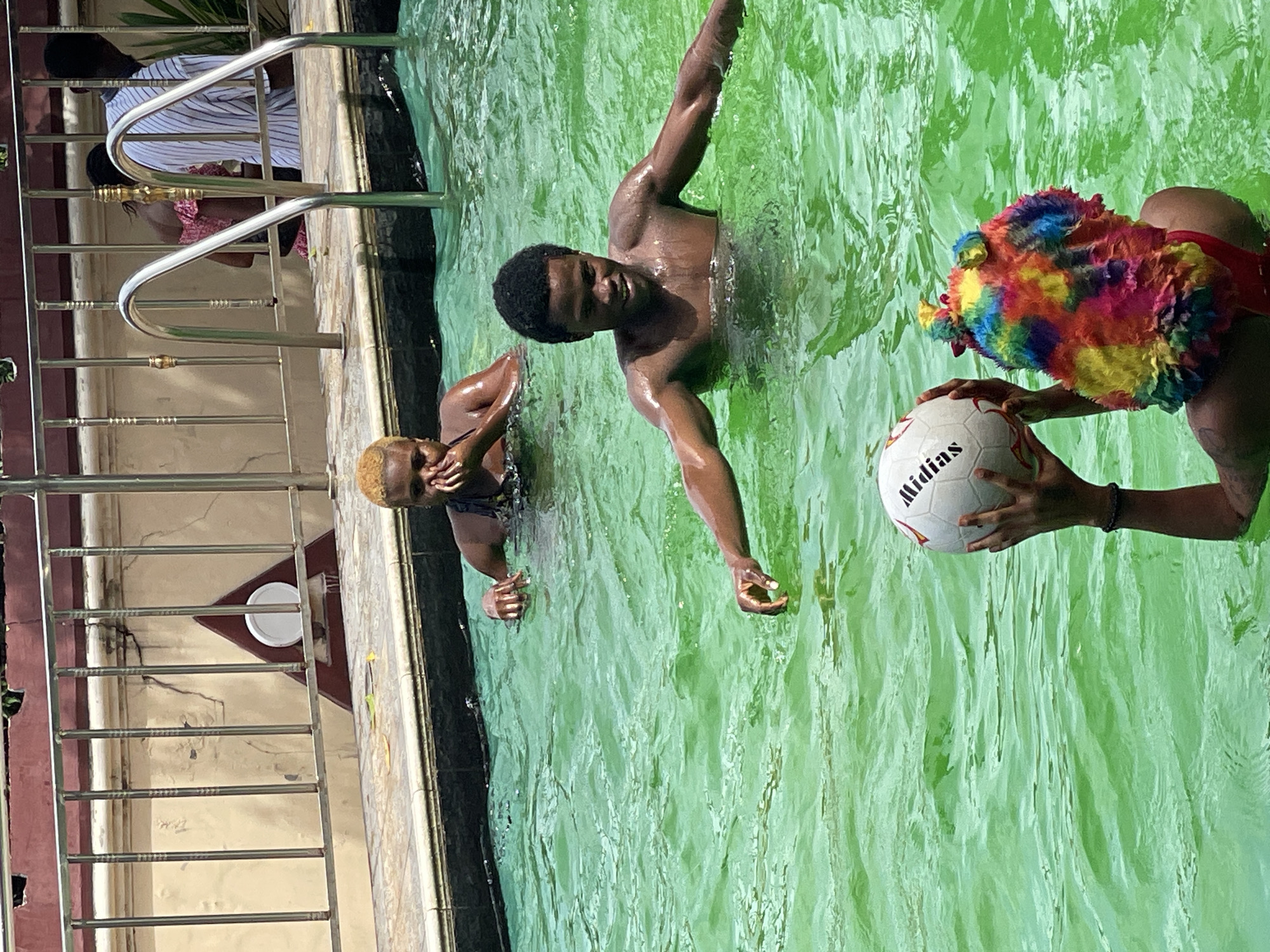 In an outdoor swimming pool filled with vibrant green water, three individuals are visible. In the foreground, a person wearing a colorful, multi-hued hat and a red bathing suit holds a white soccer ball with black lettering. To their right and slightly further back, a shirtless man with dark hair smiles broadly as he stands in the water, his arms extended outwards. In the background, a woman with blonde hair, also in the pool, rests her chin on her hands. The poolside features a tiled edge, a metal railing, and a cream-colored wall with a triangular sign.