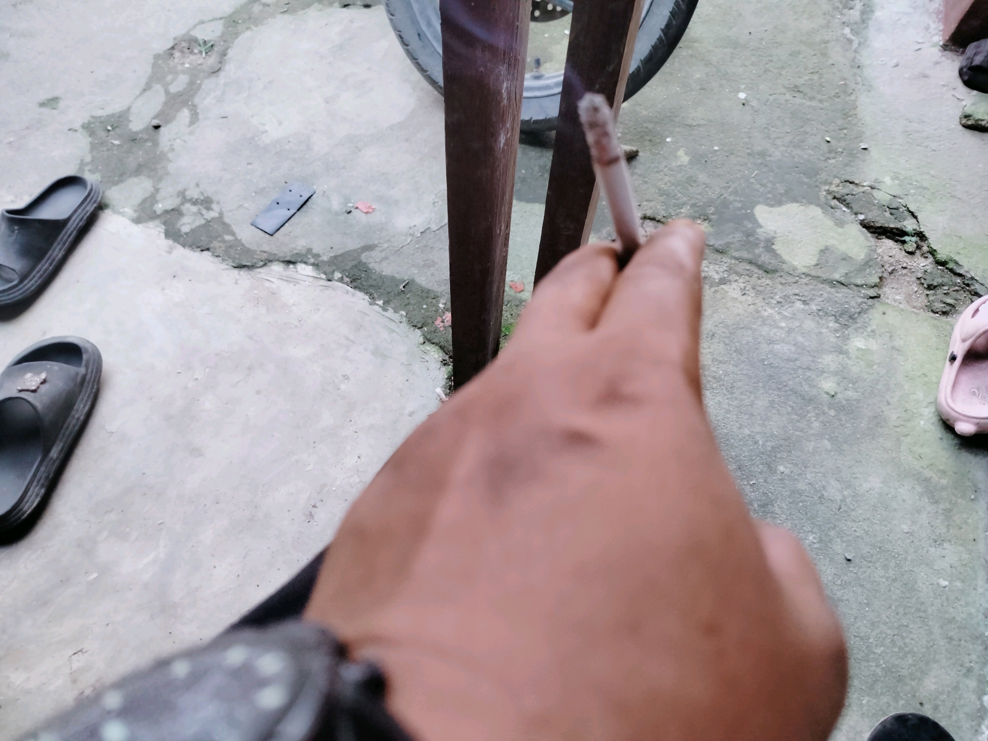 The image shows a close-up of a person's hand holding a lit cigarette. The hand is in the foreground, partially obscuring the view of the environment. In the background, there are two wooden posts, a portion of a vehicle tire, and a concrete ground surface. Scattered on the ground are a pair of black flip-flops, a dark rectangular object, and a pink sandal. The overall scene suggests an outdoor setting, possibly a casual area like a yard or walkway. The quality of the image is poor due to its low resolution and blurriness. The location, Lembahalas, Indonesia, is not directly indicated by any visual elements within the image.