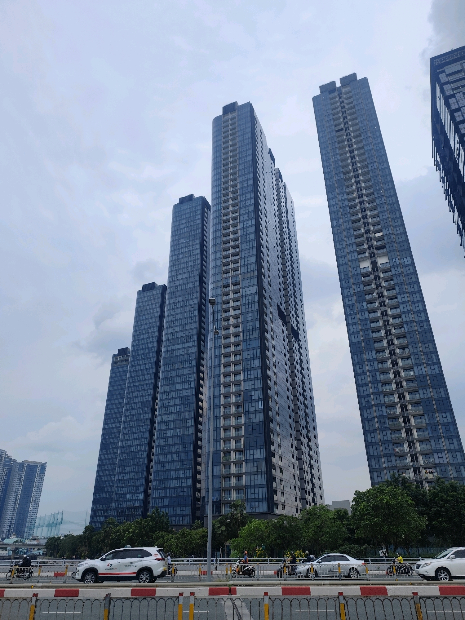 This image captures a street-level view of several modern, high-rise apartment buildings in Ho Chi Minh City, Vietnam. The buildings are characterized by their dark glass facades and numerous balconies, suggesting residential units. In the foreground, a street with a metal barrier separates the traffic from the viewer. Several vehicles, including a white SUV with commercial markings and a white sedan, are visible on the road. Motorcyclists are also present, a common mode of transportation in Vietnam. Lush green trees line the area behind the road, contrasting with the stark architecture of the skyscrapers. The sky is overcast with clouds, indicating a cloudy day. The perspective of the photo is looking upwards at the imposing structures, emphasizing their height and scale.
