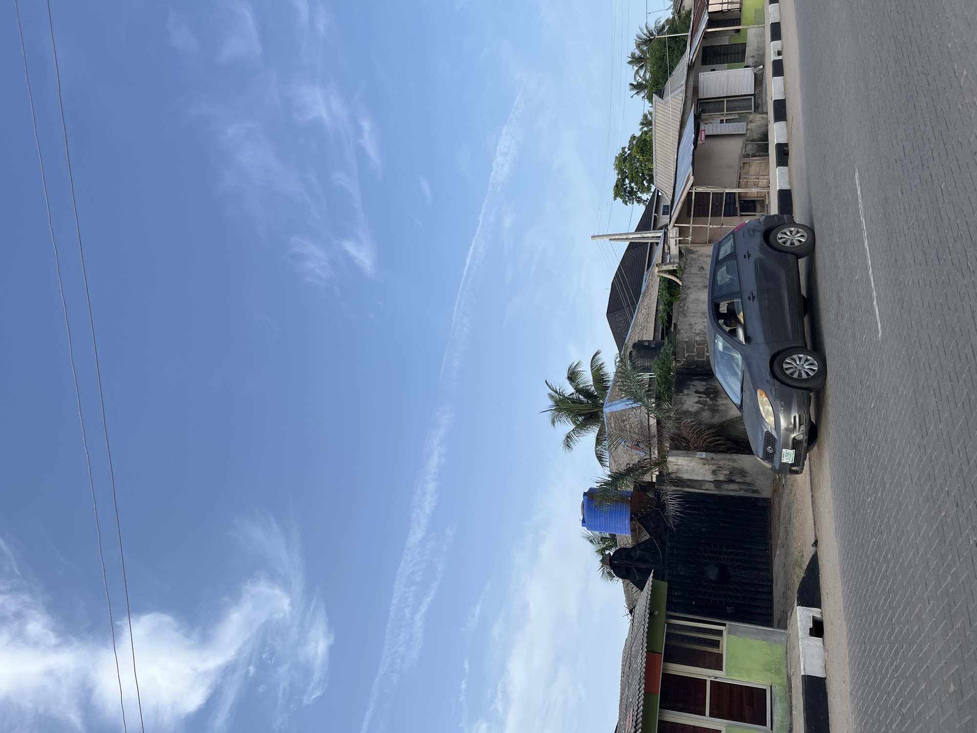 The image depicts a street scene in Badagry, Nigeria, under a clear blue sky with wispy clouds. A gray car is parked on the right side of a brick-paved road with a black and white curb. Adjacent to the road are residential buildings of varying styles and conditions, featuring corrugated metal roofs, painted walls (including shades of green and black), and some vegetation including palm trees. A blue water tank is visible atop one of the buildings. Overhead power lines are strung across the sky.
