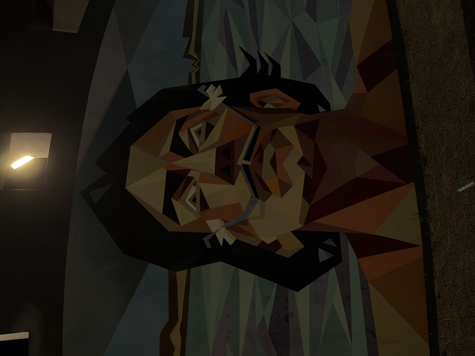 A vertically-oriented, medium shot depicts an abstract mural. The mural consists of geometric shapes, in brown, yellow, orange, blue and green hues. It contains the shapes of two faces, one on top of the other, plus the outline of a profile on the left. The mural is placed vertically against a wall which is partially visible on the left and on the right side of the image. A light fixture is visible to the left of the mural. This mural can be found at the Rubinstein Restaurant in Kazan, Russia.