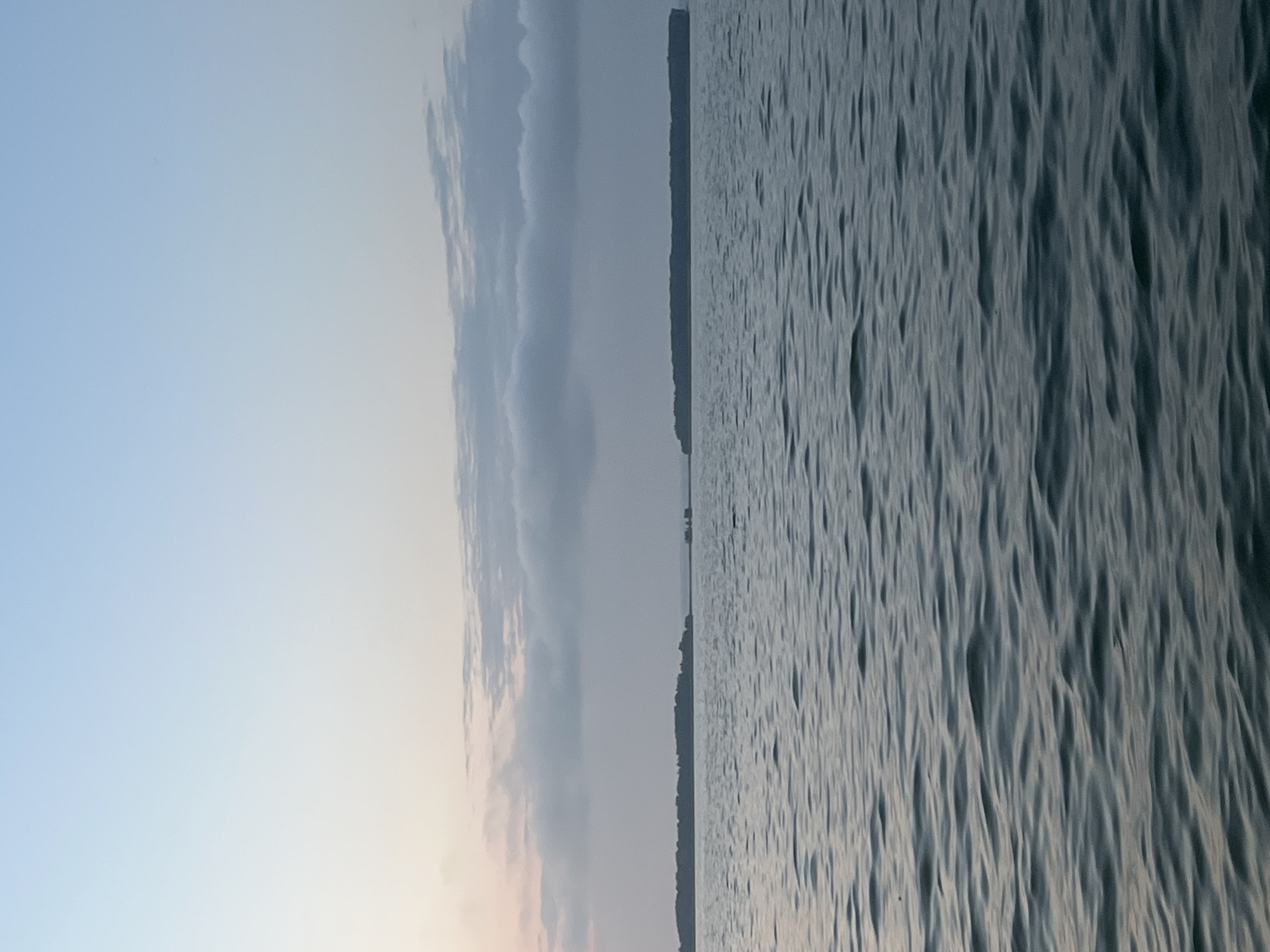 A wide shot captures a serene body of water under a soft, pale blue sky with scattered clouds. The horizon is marked by a distant, dark landmass, likely an island or peninsula, densely covered with trees. Two small, dark structures, possibly boats or buoys, are visible on the water in the distance, to the right of the landmass. The water's surface is gently rippled, reflecting the muted colors of the sky and surrounding landscape. The scene appears to be a tranquil natural setting, possibly a lake or bay, with no human presence or activity immediately apparent. This scene is not located in the city of Novopichugovo, Russia, as there is no verifiable information suggesting the image depicts this location.
