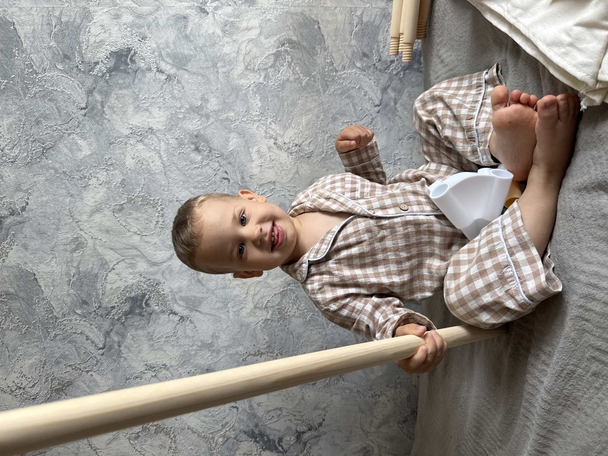 A young child, possibly male, wearing brown and white checkered pajamas, lies on a gray blanket against a textured gray and white wall. The child is holding a light-colored wooden pole. Parts of a teepee structure are visible nearby. A white plastic object rests near the child's legs. The location is presumed to be Шебунино, Russia.