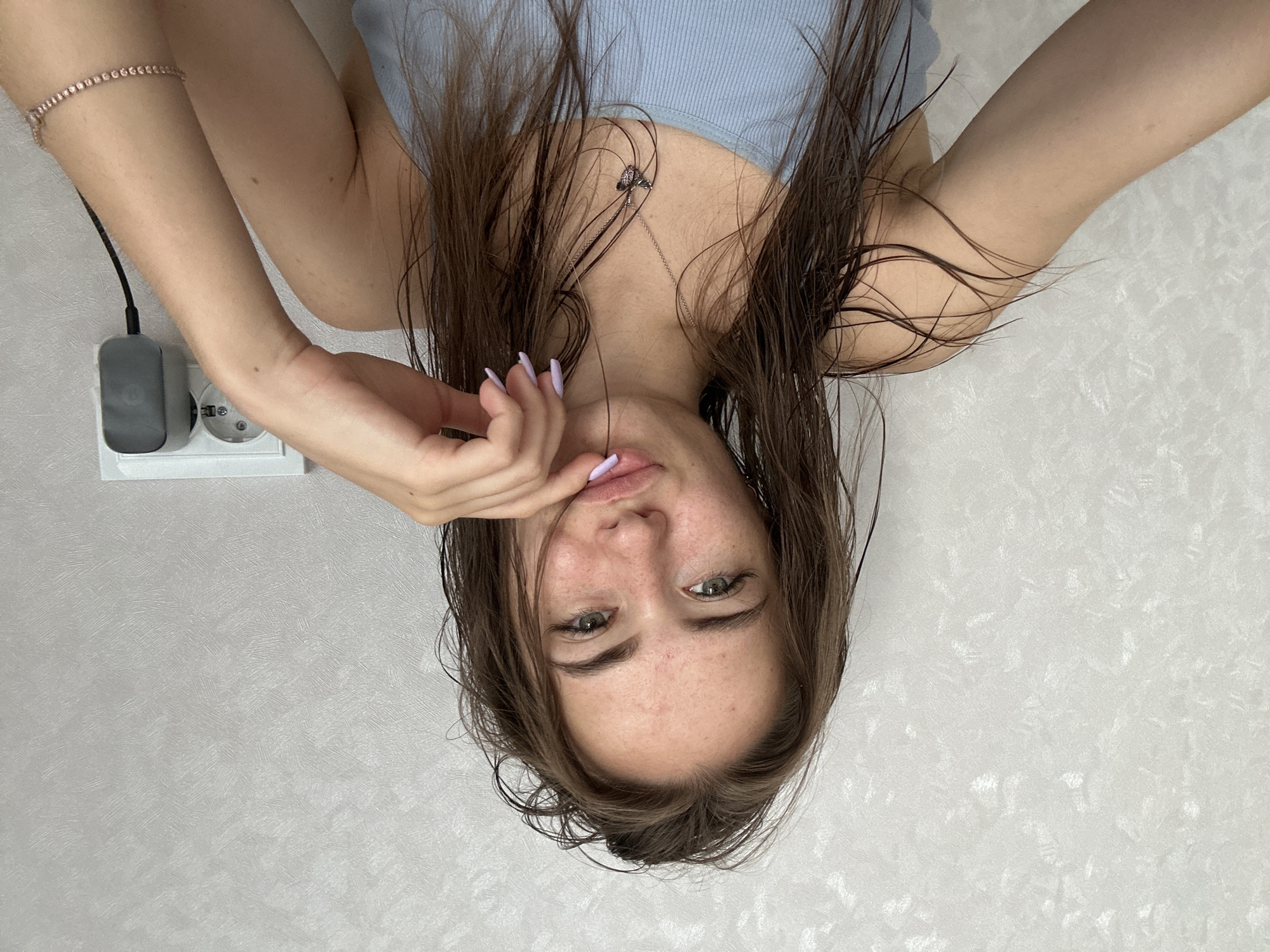 A young woman with damp, brown hair is captured in a selfie. She is wearing a light blue tank top and a delicate necklace with a butterfly pendant. Her hand is resting near her mouth, with light purple nail polish visible. In the background, a white wall with a textured pattern is visible, along with a white electrical outlet with a gray adapter plugged in. The overall setting appears to be indoors, in an unremarkable room.