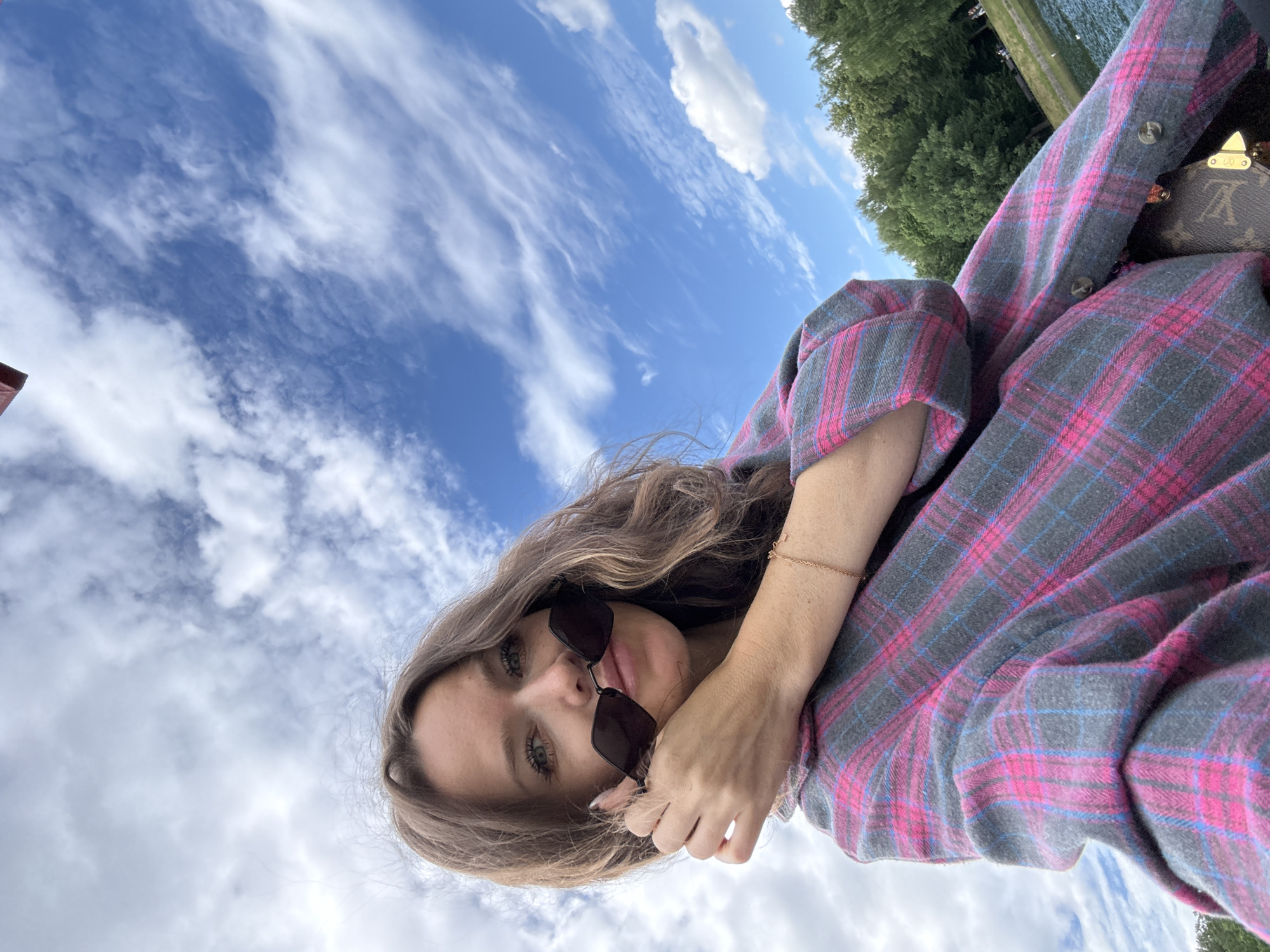 A woman is taking a selfie outdoors with a blue sky and clouds in the background. She is wearing a pink and gray plaid shirt and sunglasses, which she is holding up to her face. Her hand is near her mouth, and she has a bracelet on her wrist. Behind her, there are trees and a body of water. The image appears to have been taken in Moscow, Russia.
