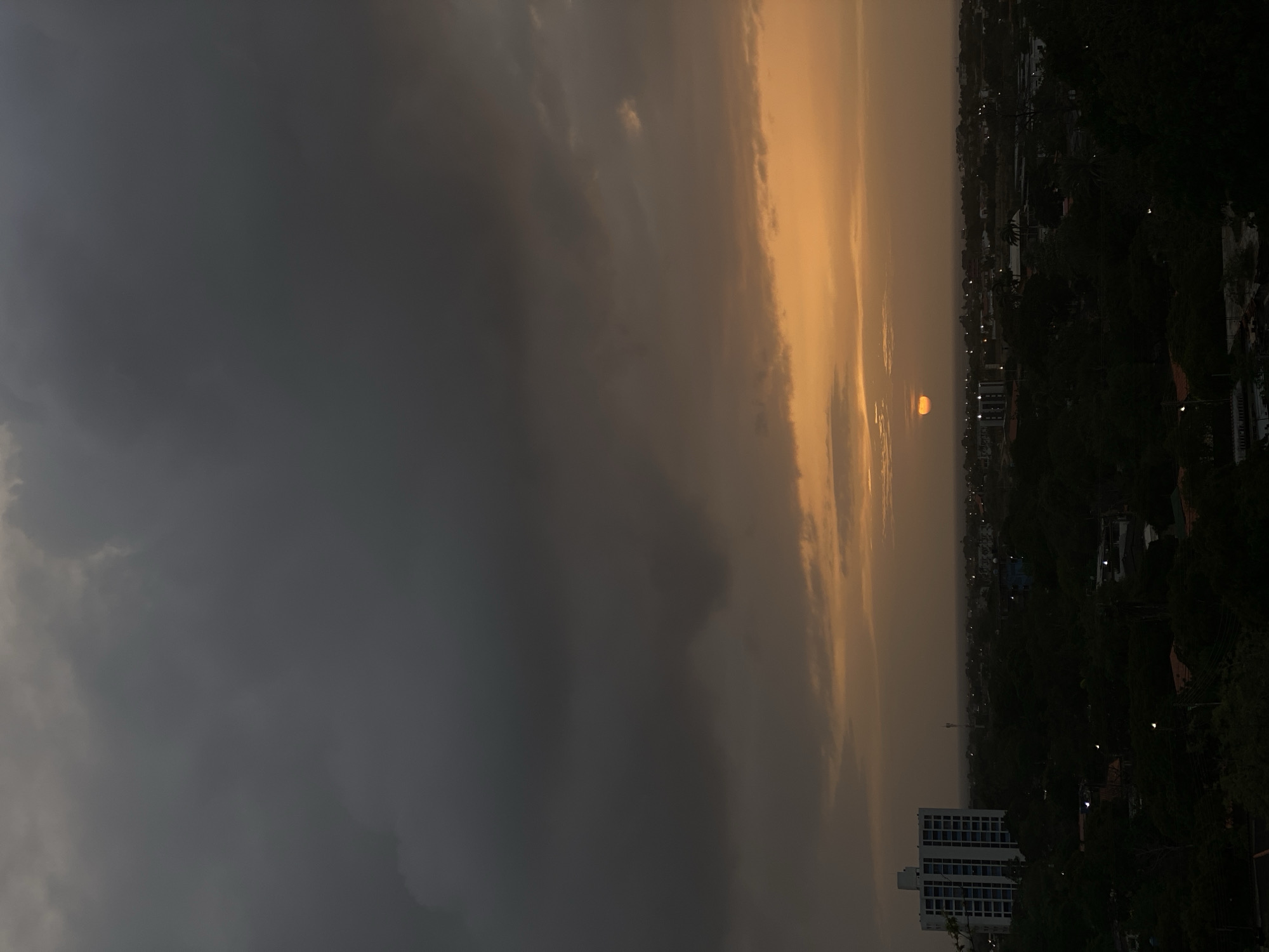 This is an aerial, vertical, medium shot of a sunset over the city of Juan Griego, Venezuela. Dark, ominous clouds fill the upper half of the frame, with a sliver of orange sunlight breaking through near the horizon, where the sun itself is partially visible as a small orange disk. Below the sky, the city stretches out, mostly obscured by the dark silhouettes of trees. A tall, white building with blue accents stands out prominently on the left side of the image. The scene is characterized by low light and a dramatic, cloudy sky.